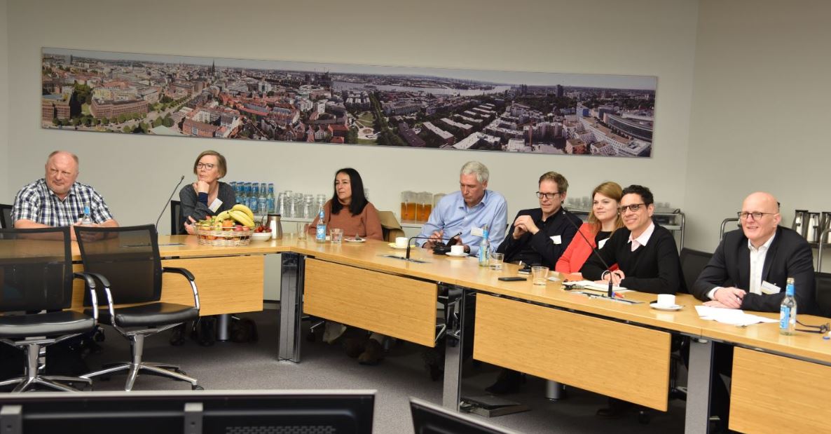 8 Menschen sitzen an Tischen in einem Konferenzraum. Im Hintergrund eine Panoramaansicht von Hamburg.