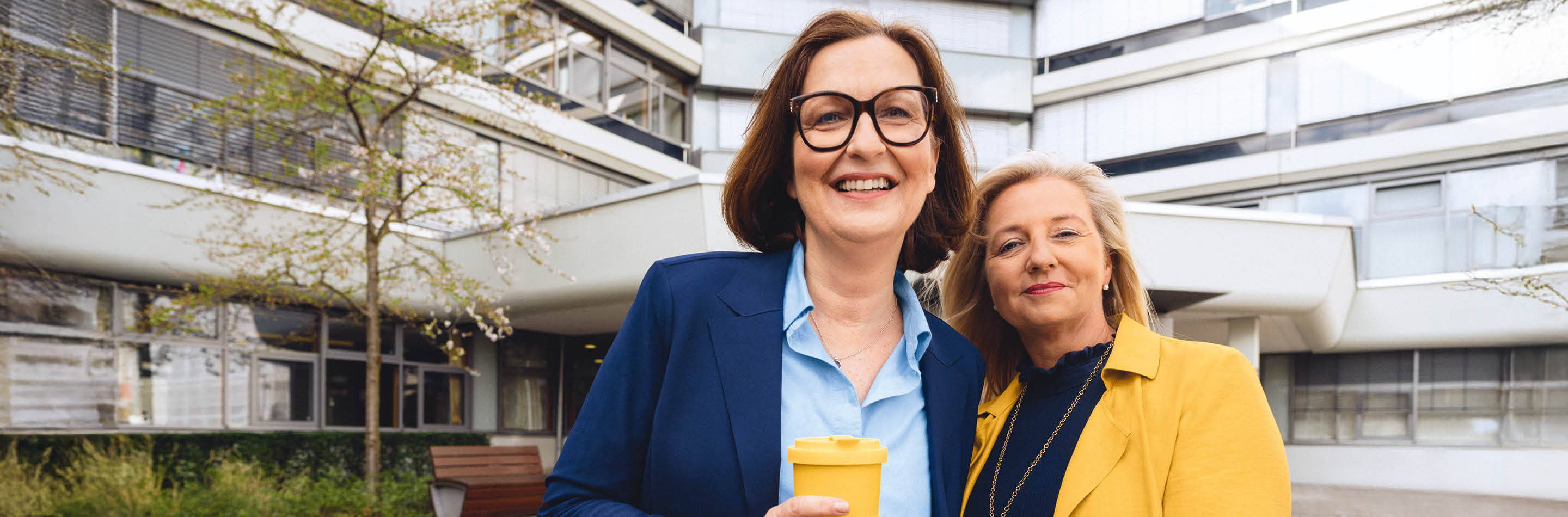 1000 UNDEIN GRUND – Deine Idee von Karriere: mehr bewirken. Zwei Frauen stehen lächelnd vor dem Gebäude der Hauptverwaltung der BGW in Hamburg.