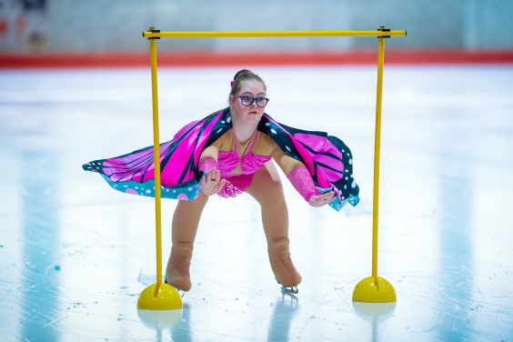 Junge Eislaufkünstlerin mit pinkem Umhang fährt unter Hindernis hindurch