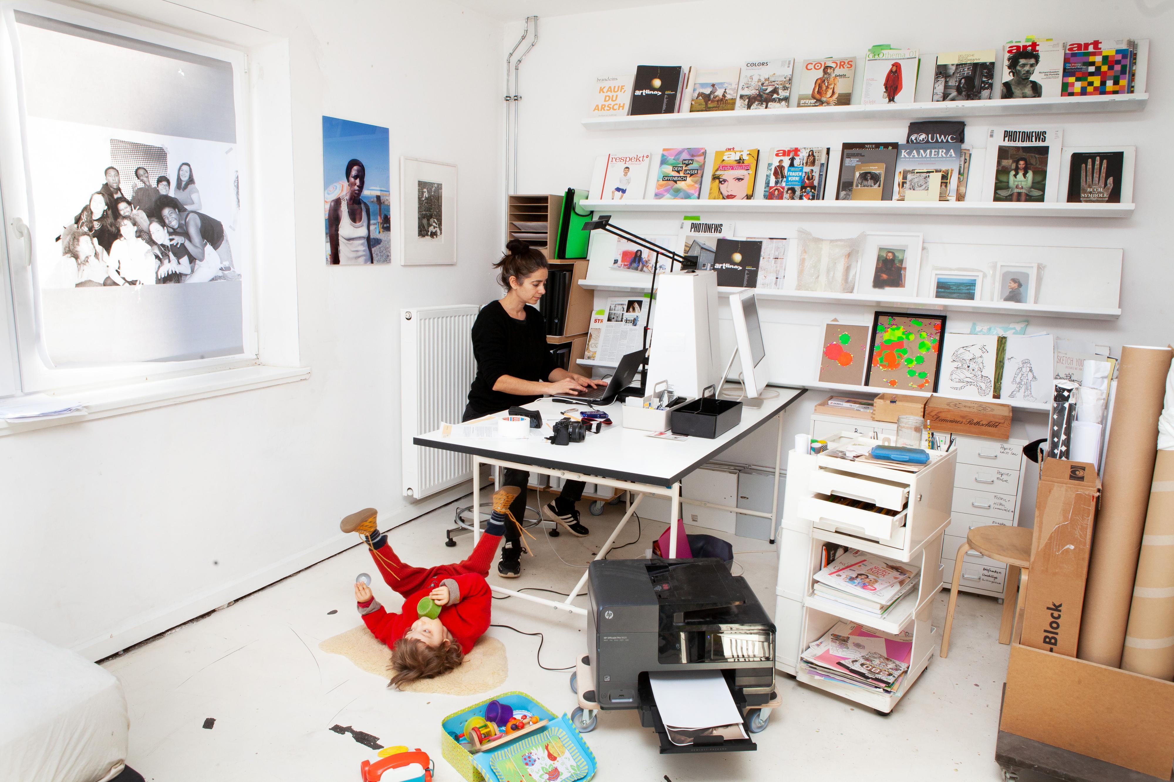 Eine Frau steht an einem Tisch und arbeitet am Computer. Vor dem Tisch liegt ein Kleinkind auf dem Boden und spielt. Im Raum herrscht Unordnung.
