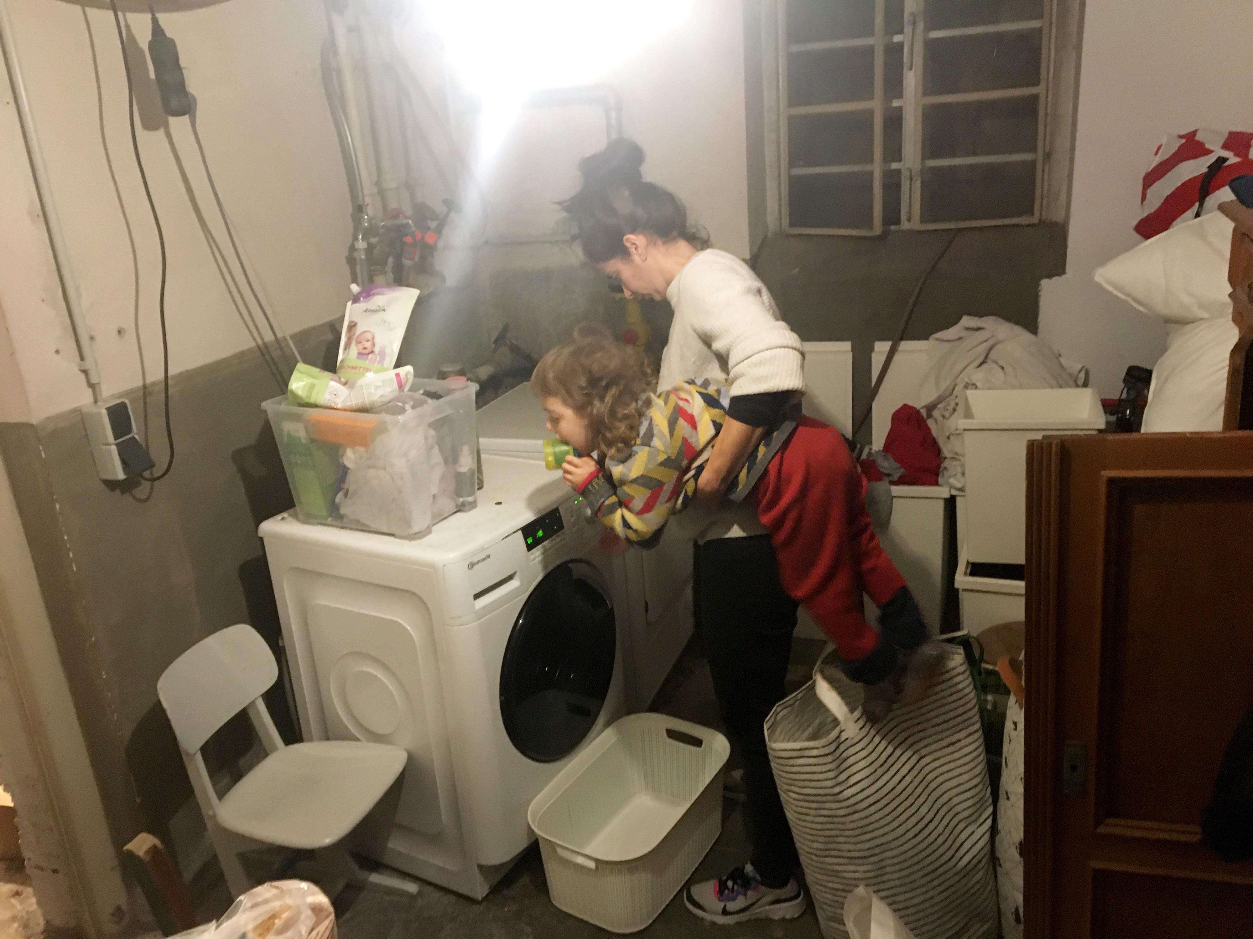 Eine junge Frau steht vor einer Waschmaschine in einem Kellerraum. Sie hat ein Kleinkind unter dem Arm. Im Keller stehen und liegen überall Gegenstände herum.
