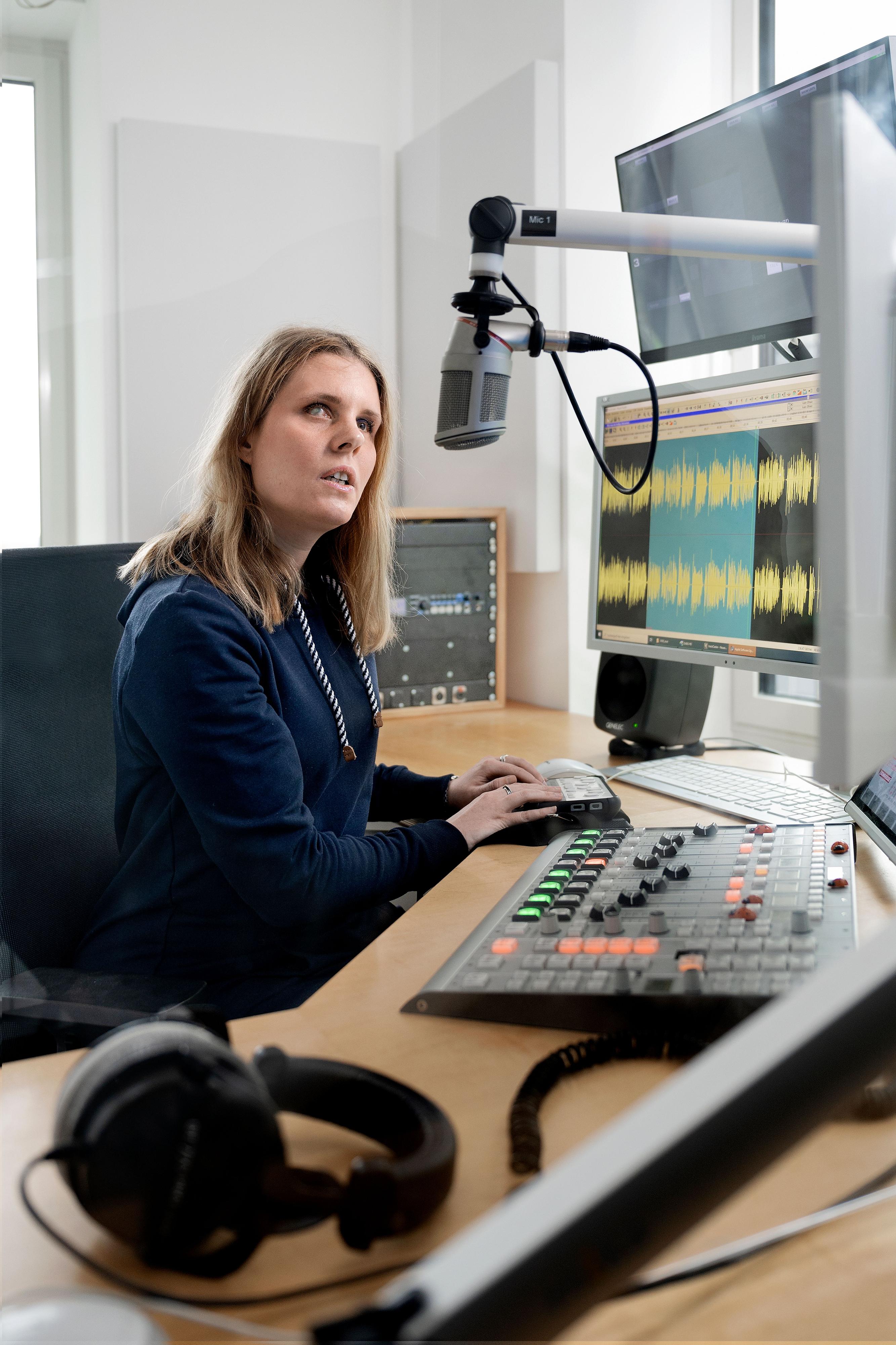 Eine blinde junge Frau sitzt in einem Radiostudio und spricht in ein Mikrofon. Sie hat die Hände auf einem Gerät abgelegt. 