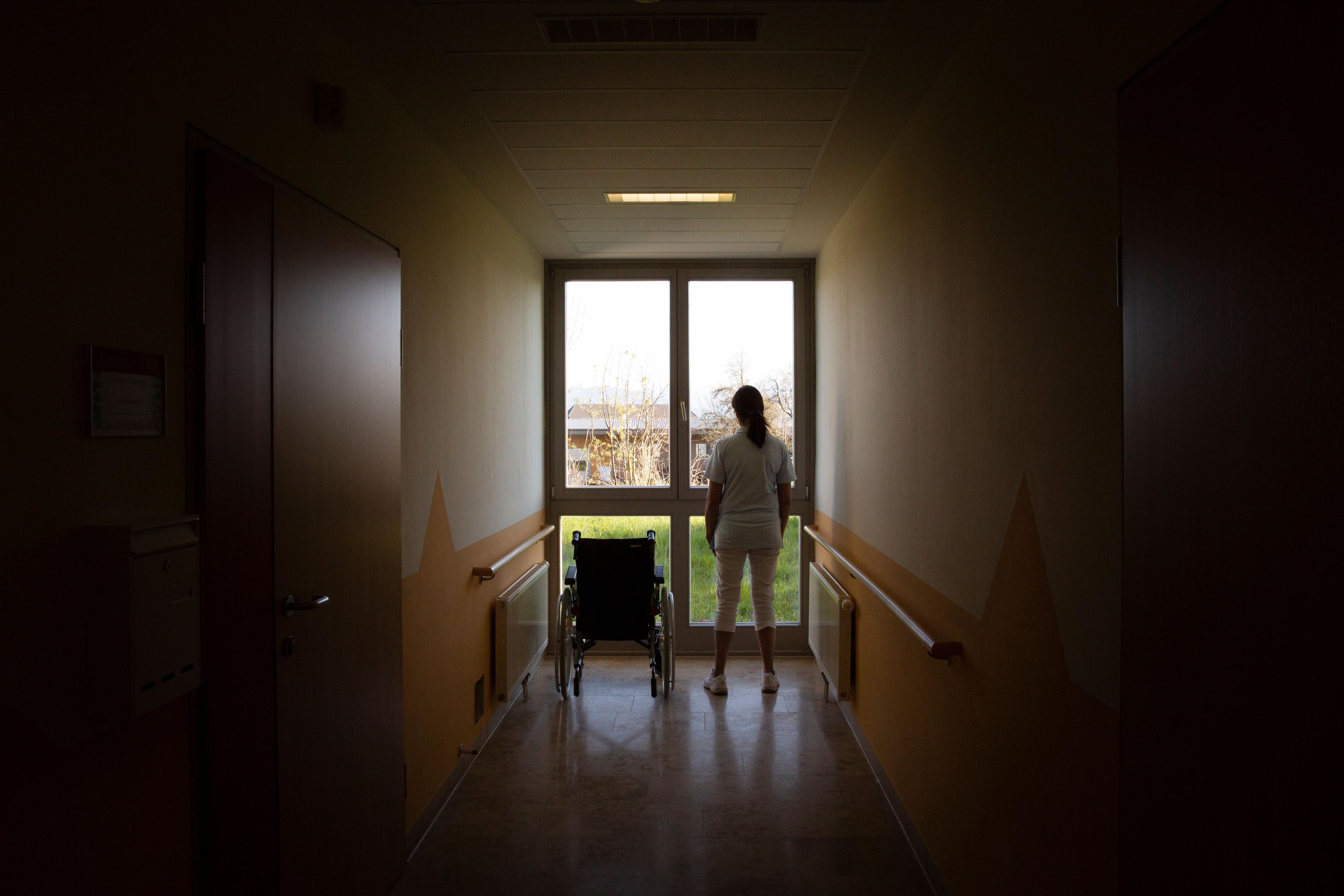 Eine Pflegerin steht neben einem leeren Rollstuhl vor einem Fenster.