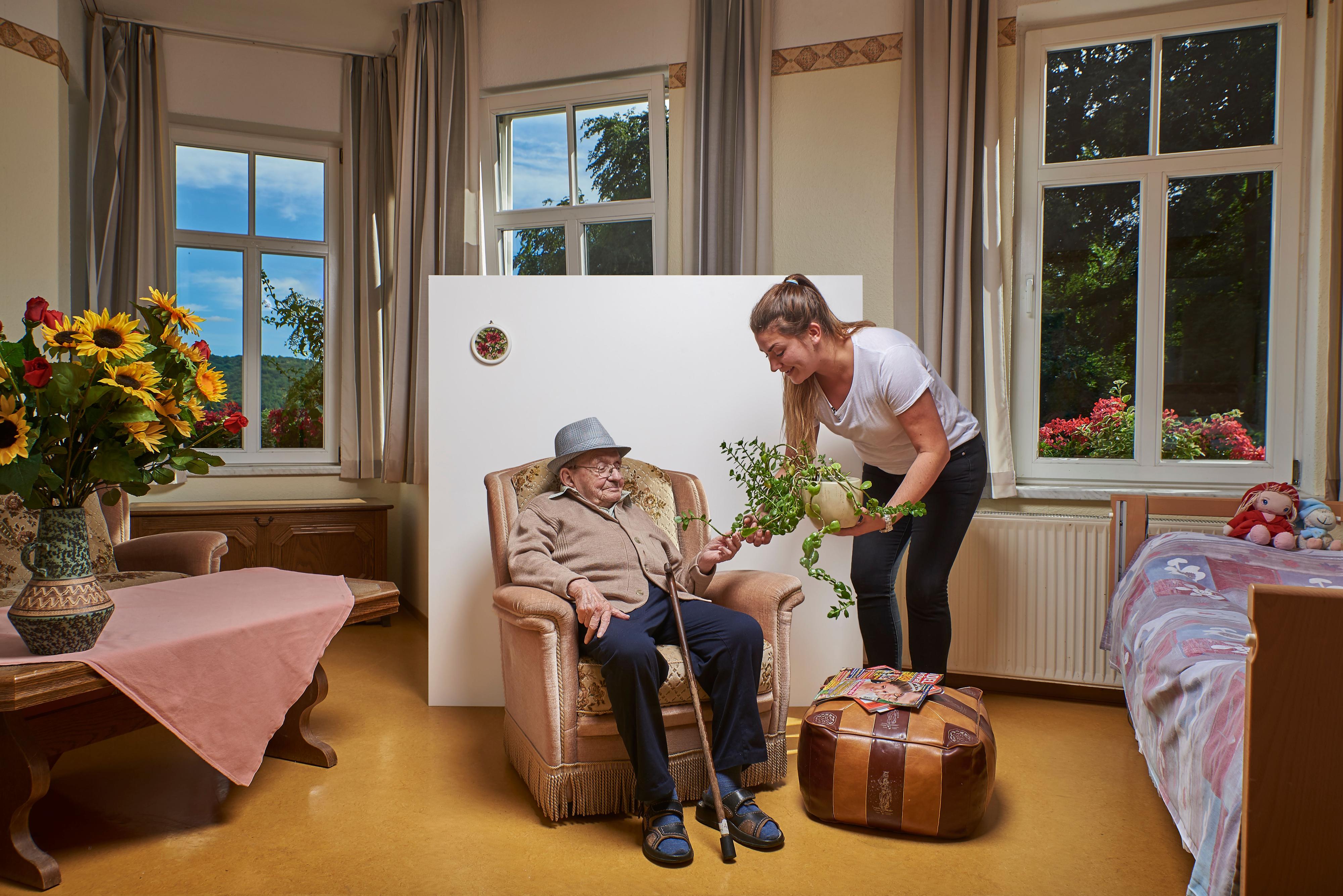 Eine Pflegerin mit einem Blumentopf in der Hand steht vor einem älteren Mann in einem Sessel.