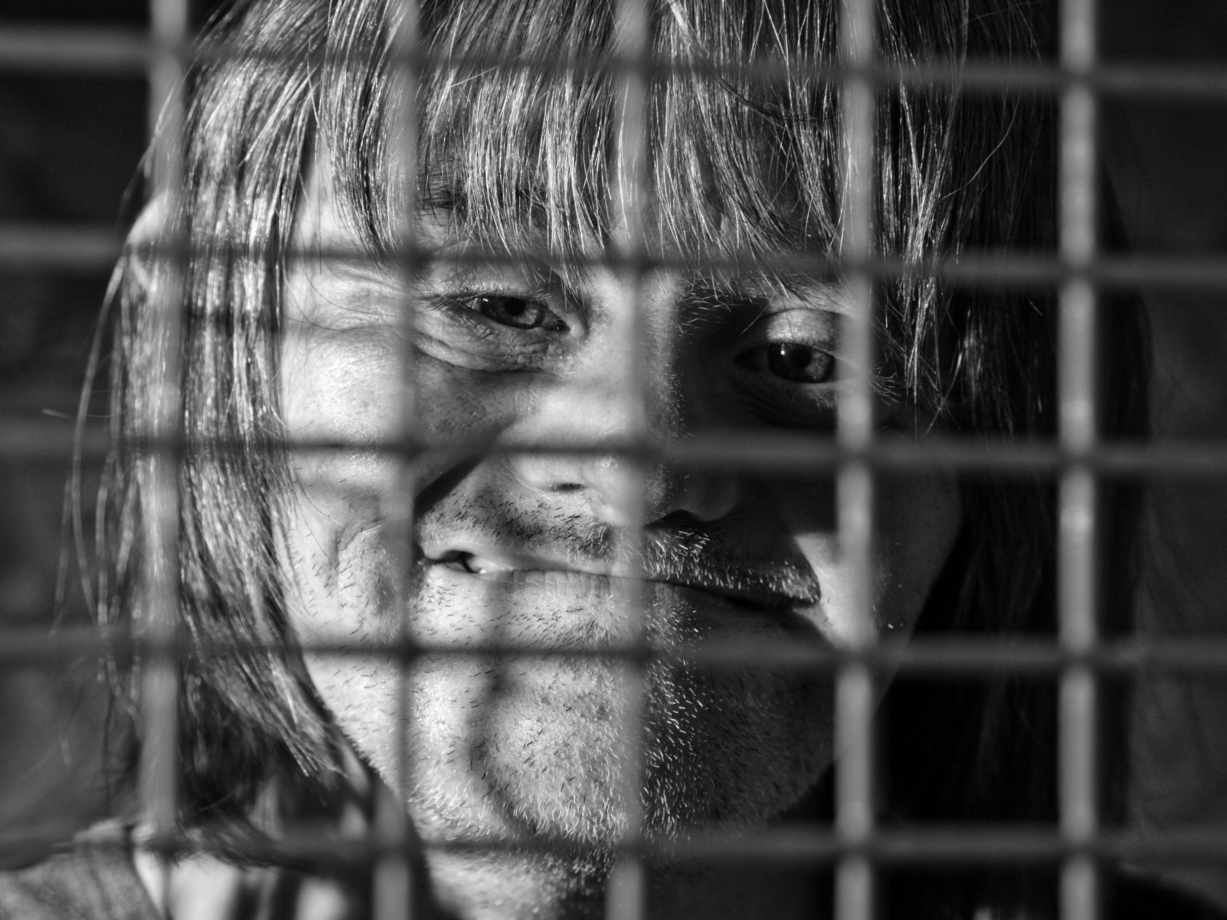 Ein Mann - mit phänotypischen Zügen der Trisomie 21 - lächelt, wobei er hinter einem Gitter zu sehen ist. 