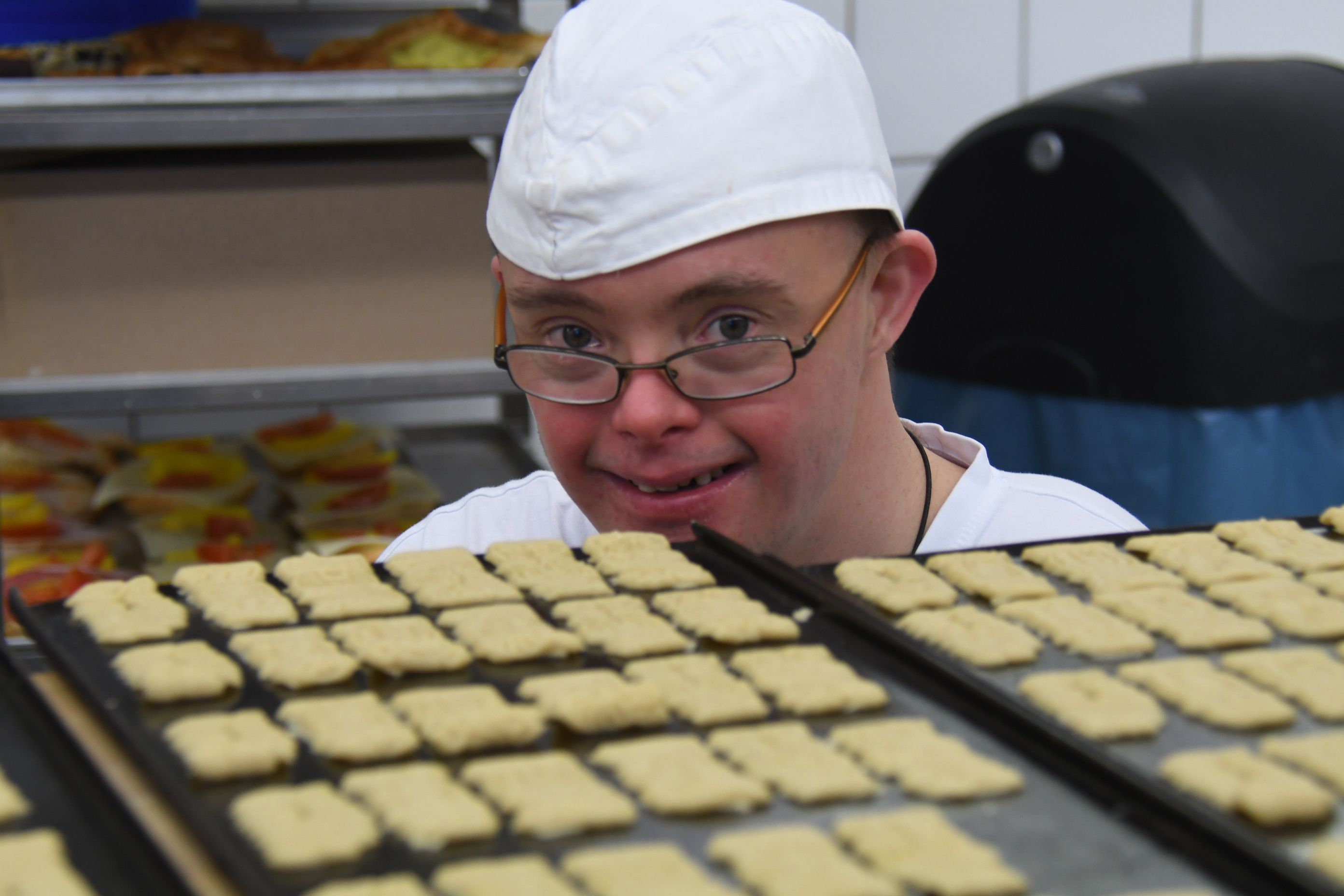 Ein junger Mann mit Behinderung hinter zwei Backblechen voller Kekse. Er trägt eine Bäckermütze und schaut über seinen oberen Brillenrand in die Kamera.