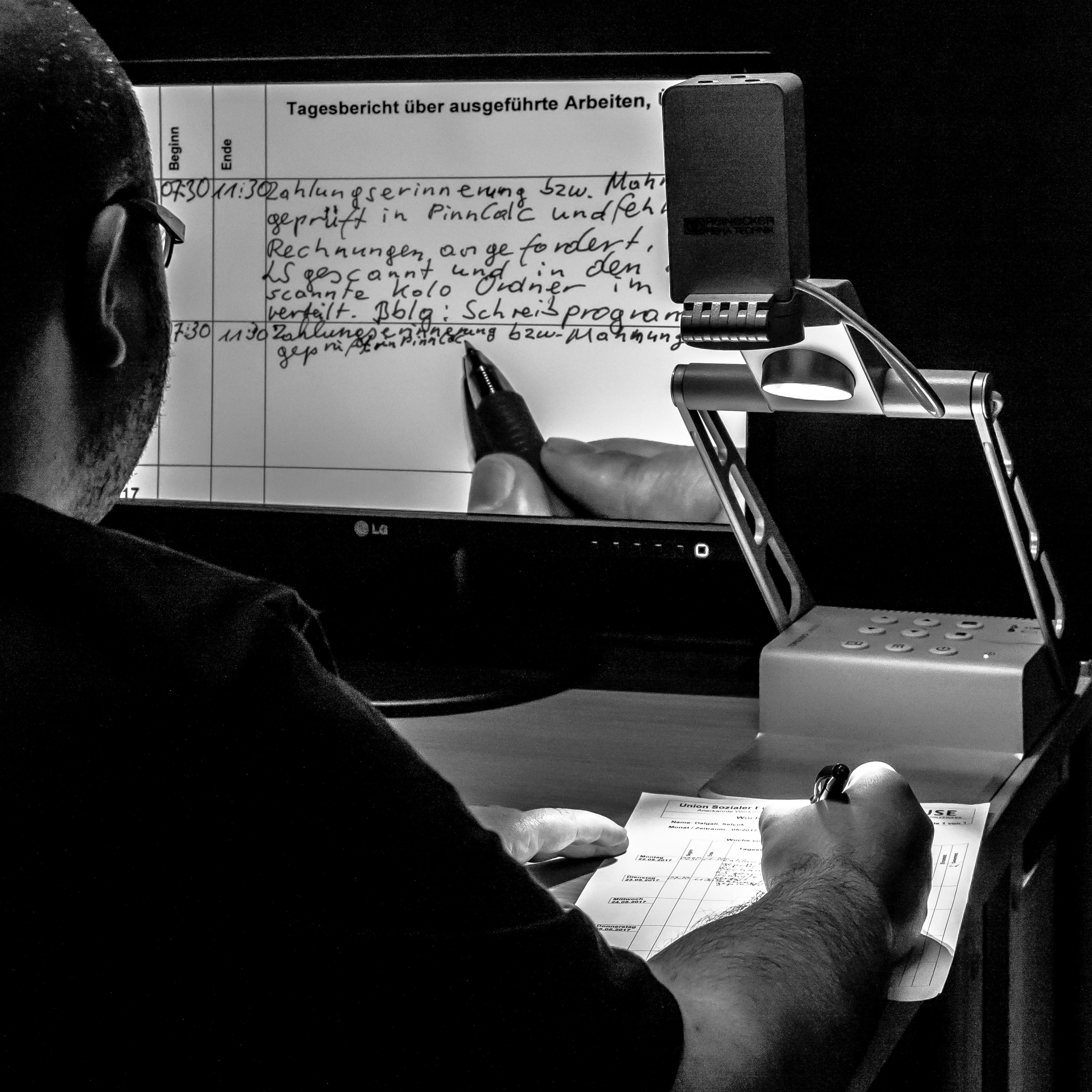 Ein Mann hält einen Zettel unter eine Lampe und schaut auf einen Monitor.