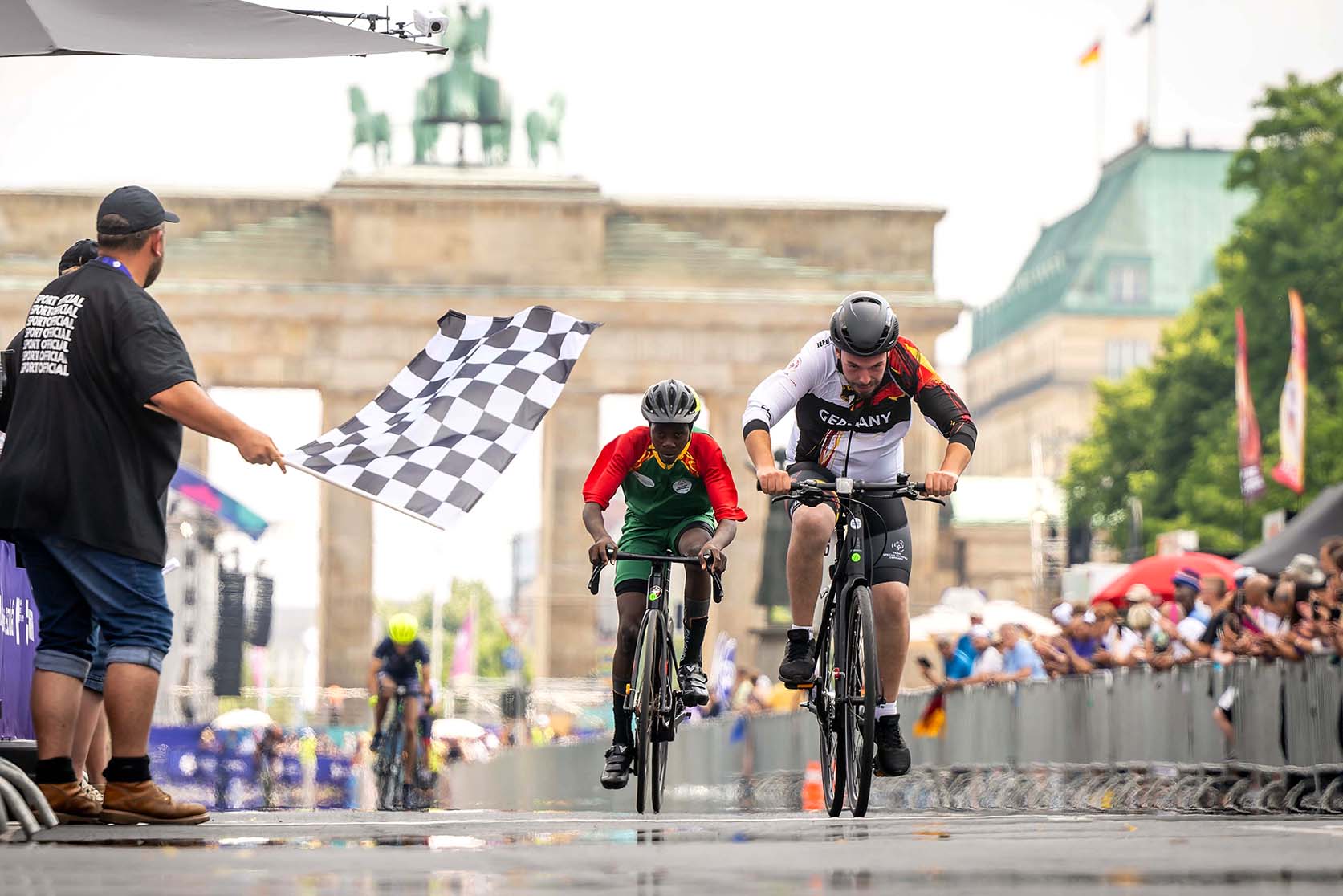 Zwei Radrennfahrer auf der Rennstrecke. Links ein Mann mit Zielflagge, rechts Zuschauende am Rand der Straße. Im Hintergrund das Brandenburger Tor.