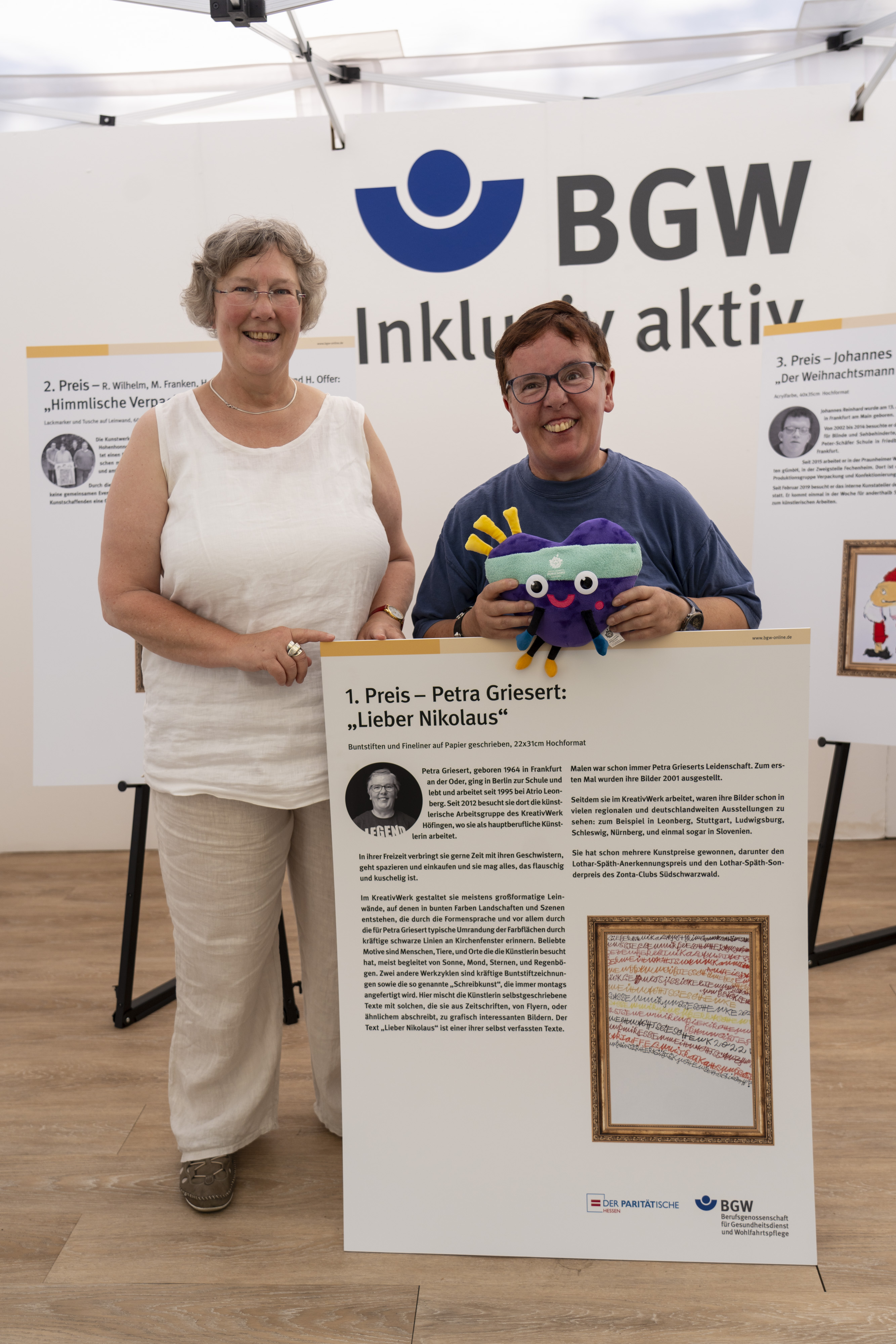  Silvia Thimm zusammen mit der Gewinnerin Petra Griesert. Die Gewinnerin hält das Maskotchen Unity in der Hand und steht hinter einer Tafel mit ihrem Bild und Text dazu am BGW-Stand.