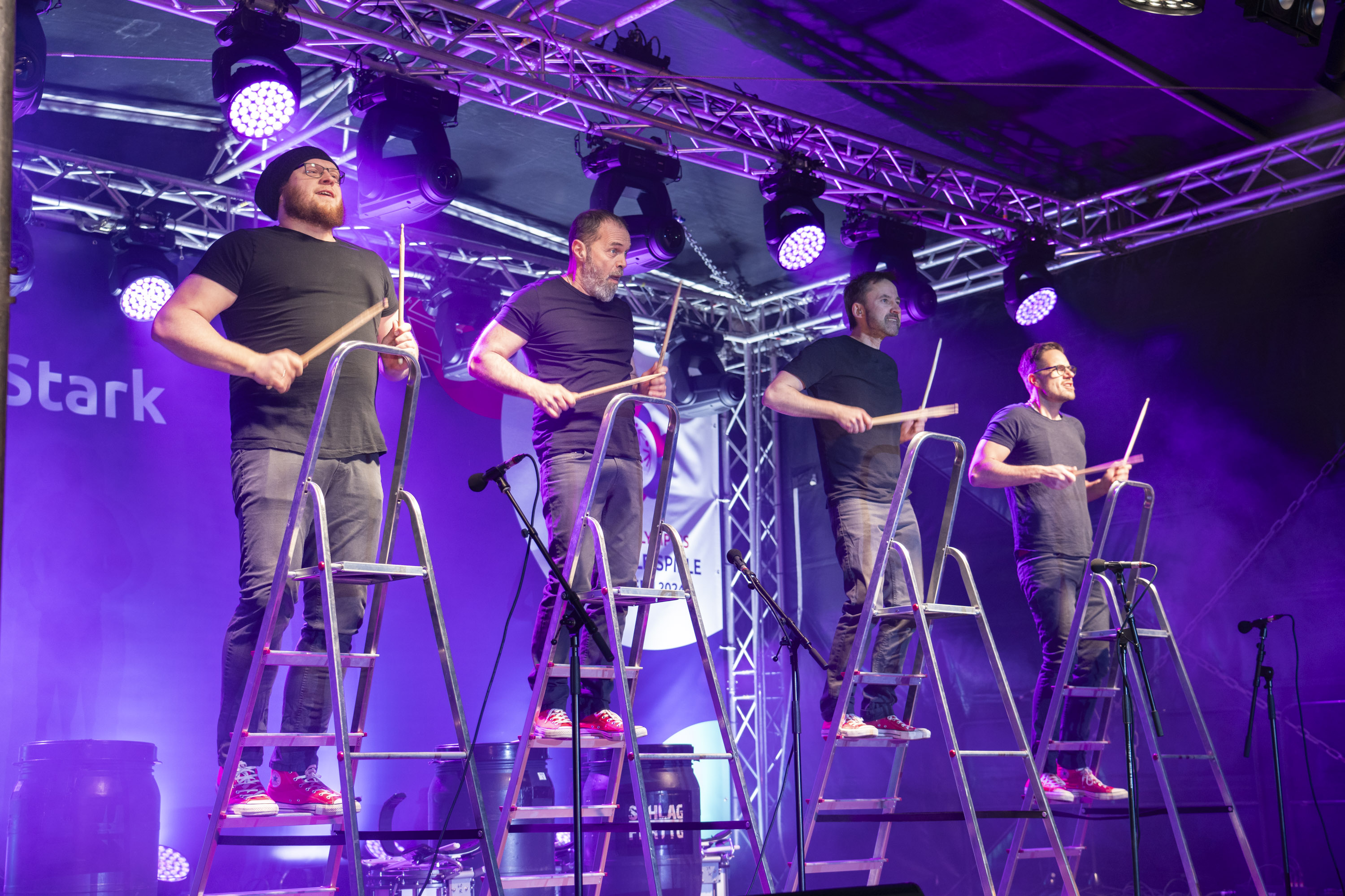 4 Trommler stehen auf Leitern auf einer Bühne und Trommeln mit Stöcken auf die Leitern.
