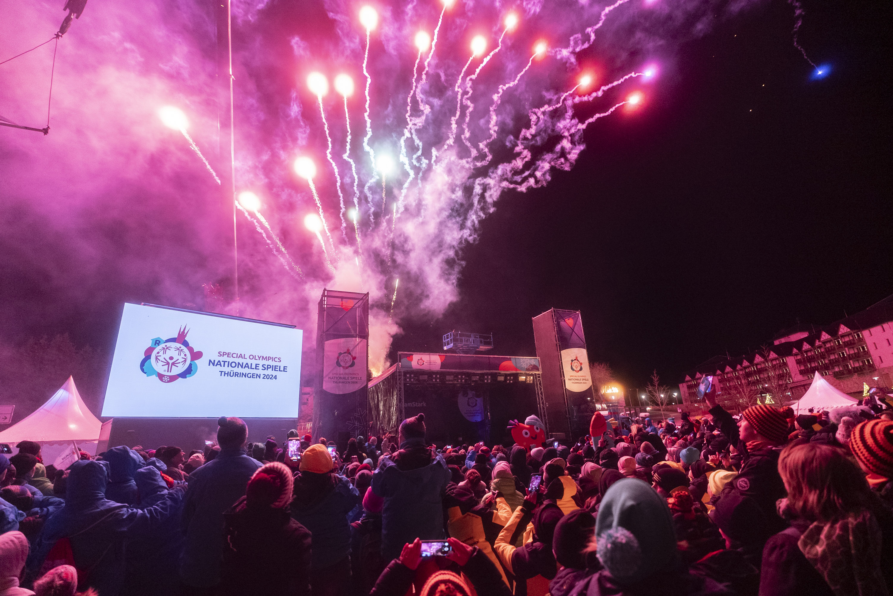 Ein Feuerwerk im Himmel hinter einer Bühne, davor eine Menschenmenge und eine Leinwand mit dem Logo der Spiele