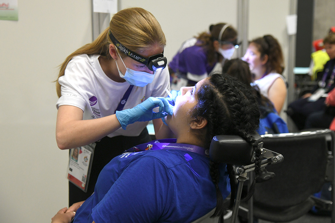 Eine Zahnärztin mit Mundschutz, Handschuhen und Stirnlampe leuchtet einer Teilnehmerin in den geöffneten Mund.