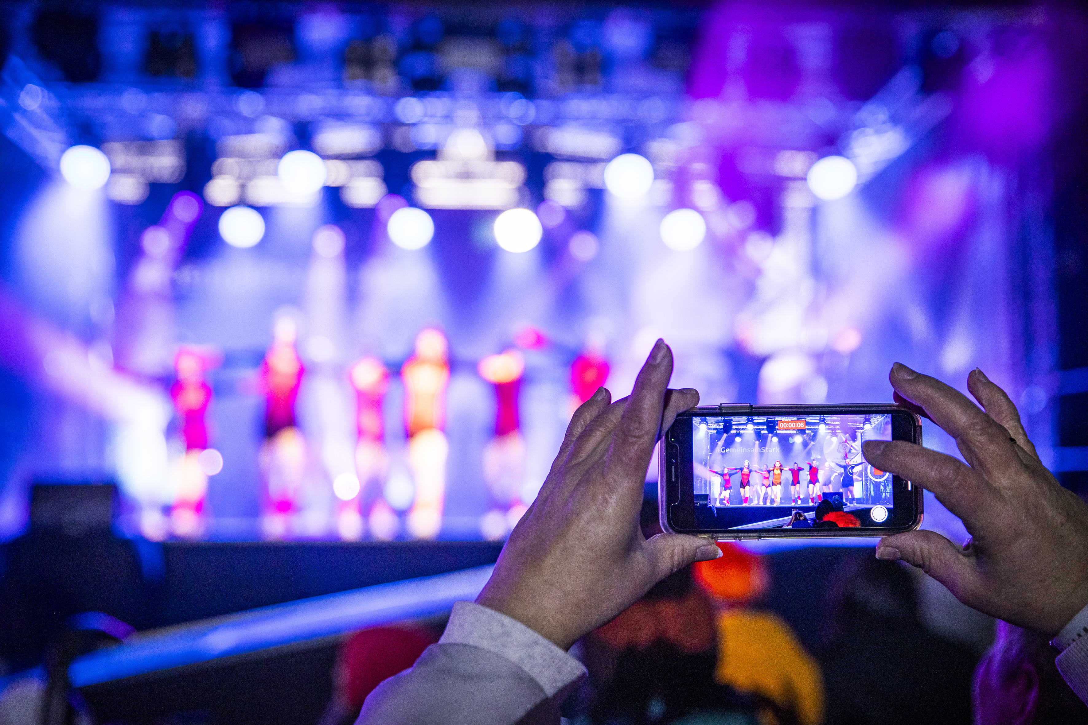 Im Hintergrund verschwommen Tänzerinnen und Tänzer auf der Bühne, im Vordergrund zwei Hände, die ein Handy halten, das filmt.