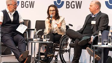 2 Männer und eine Frau auf einem Podium. Der linke hat eine Behinderung an beiden Armen. Die Frau in der Mitte sitzt im Rollstuhl.