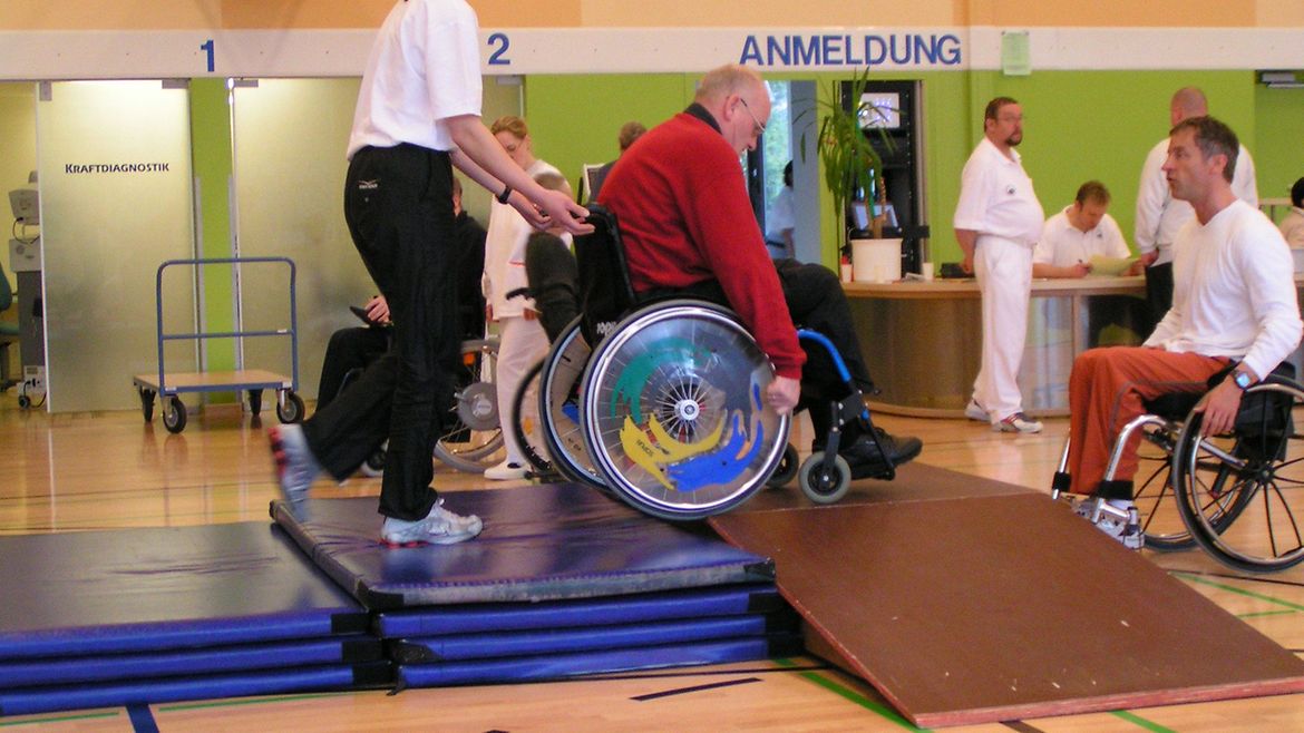 Trainerin liftet einen Rollstuhlnutzer über ein Hindernis.