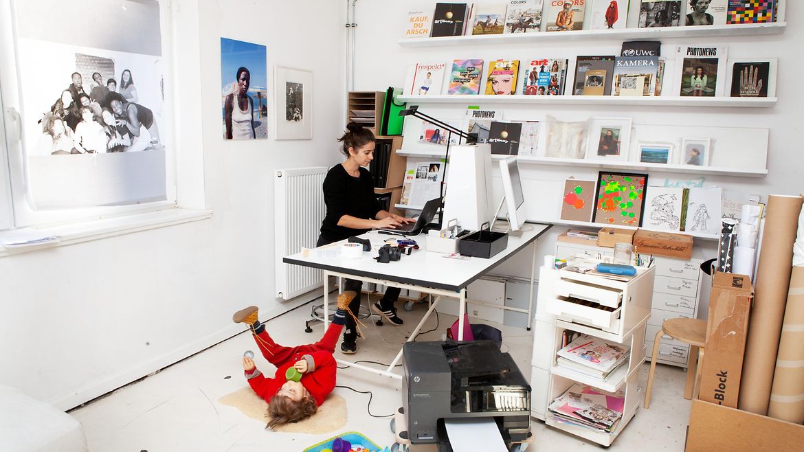 Eine Frau steht an einem Tisch und arbeitet am Computer. Vor dem Tisch liegt ein Kleinkind auf dem Boden und spielt. Im Raum herrscht Unordnung.