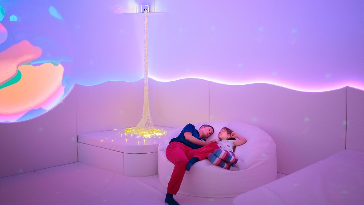 Eine Frau und ein Kind liegen in einem farbig beleuteten Raum auf einem großen runden Kissen und entspannen sich.