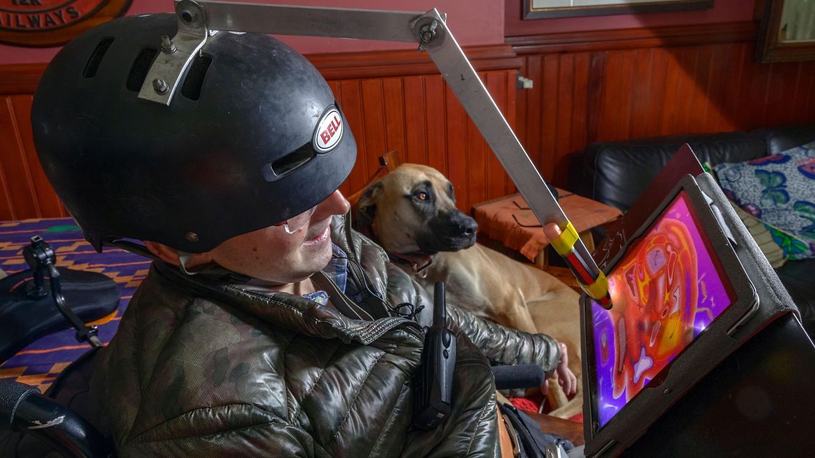 Ein Mann mit Helm auf dem Kopf. Der Helm hat eine Vorrichtung mit einem Stift, der Farben auf einen Bildschirm malt. Im Hintergrund liegt ein großer brauner Hund.