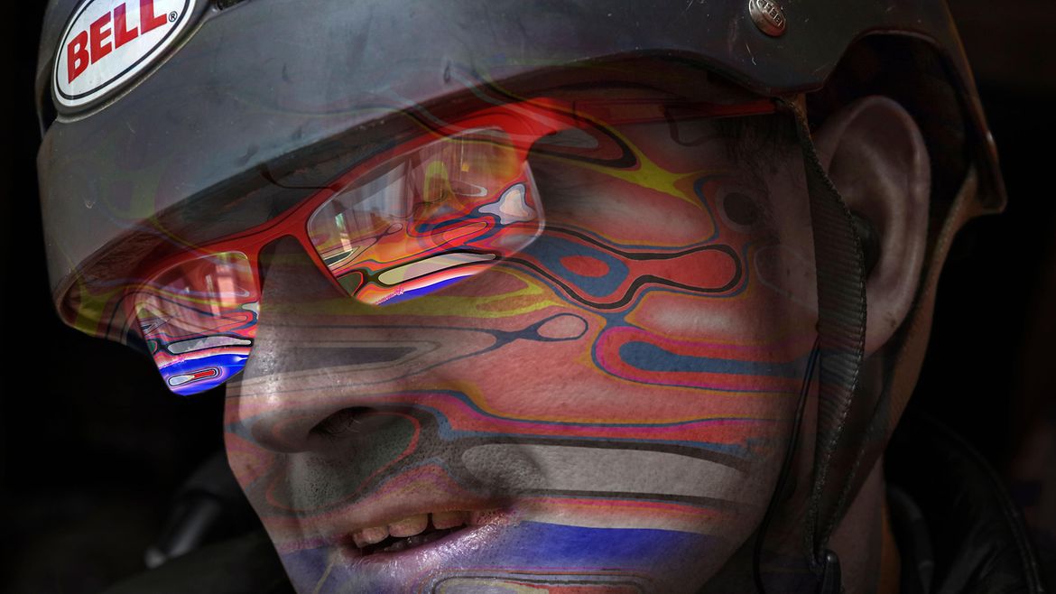 Das Gesicht eines Mannes mit Helm auf dem Kofp und Brille. Auf seinem Gesicht spiegelt sich ein buntes Gemälde.
