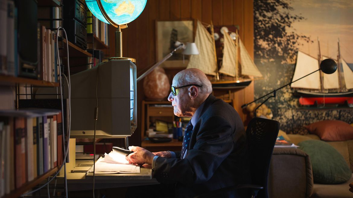 Ein alter Mann mit großer Brille sitzt in einem Wohnzimmer ganz nah vor einem Monitor und blättert in Unterlagen. Auf dem Monitor steht eine beleuchtete Weltkugel. Der Lichtschein trifft sein Gesicht.