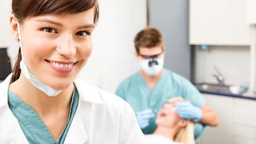 Zahnmedizinische Fachangestellte. Im Hintergrund behandelt ein Zahnarzt eine Patientin.
