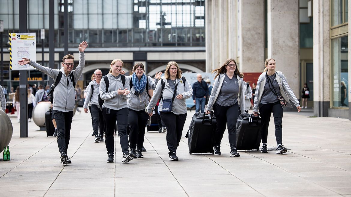 Eine Gruppe Sportlerinnen und Sportler in grau-schwarzem Outfit mit Koffern vor einem Bahnhof