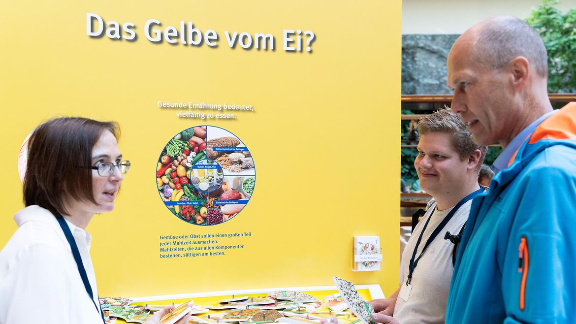 Zwei Personen informieren sich an einem Stand zum Thema Ernährung mit dem Titel "Das Gelbe vom Ei" bei einer Frau.