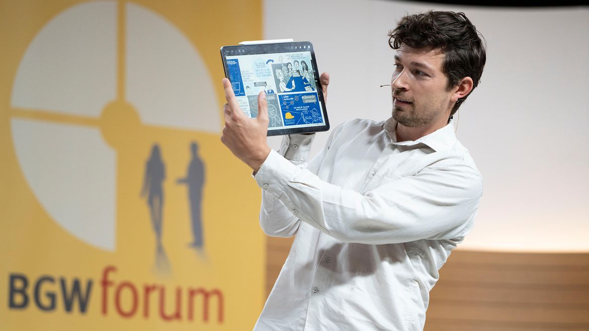 Graphic Recorder Manuel Recker zeigt auf der Bühne ein Tablet mit seinen Zeichnungen.