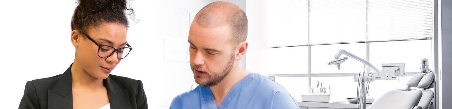 Junge Frau mit tablet und junger Mann in blauer Pflegekleidung mit Klemmbrett schauen zusammen auf den Bildschirm des tablets. Im Hintergrund: Behandlungszimmer einer Zahnarztpraxis.