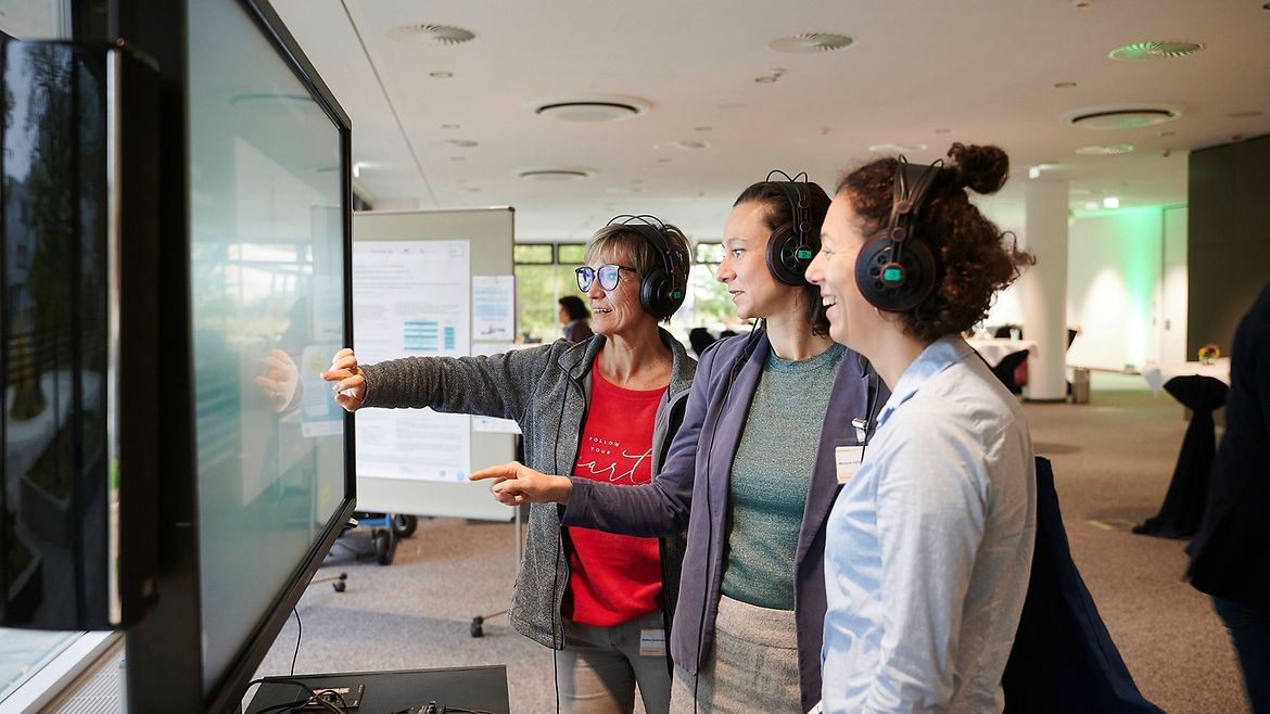 Drei Frauen stehen mit Kopfhörern auf den Ohren vor einem großen Bildschirm, eine berührt den Bildschirm