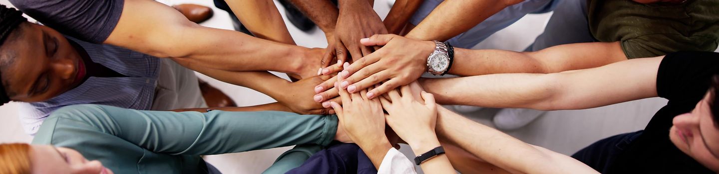Gruppe von Menschen mit verschiedenen Hautfarben stehen in einem Kreis. In der Mitte halten sie ihre ausgestreckten Hände übereinander.