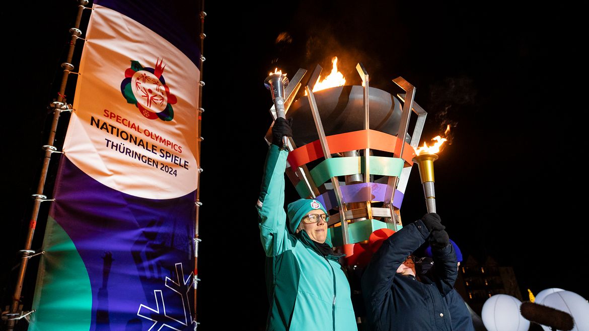 Zwei Sportlerinnen stehen vor der entzündeten Flamme und halten beide Fackeln in der Hand. Links daneben ein Banner mit dem Logo der Spiele.