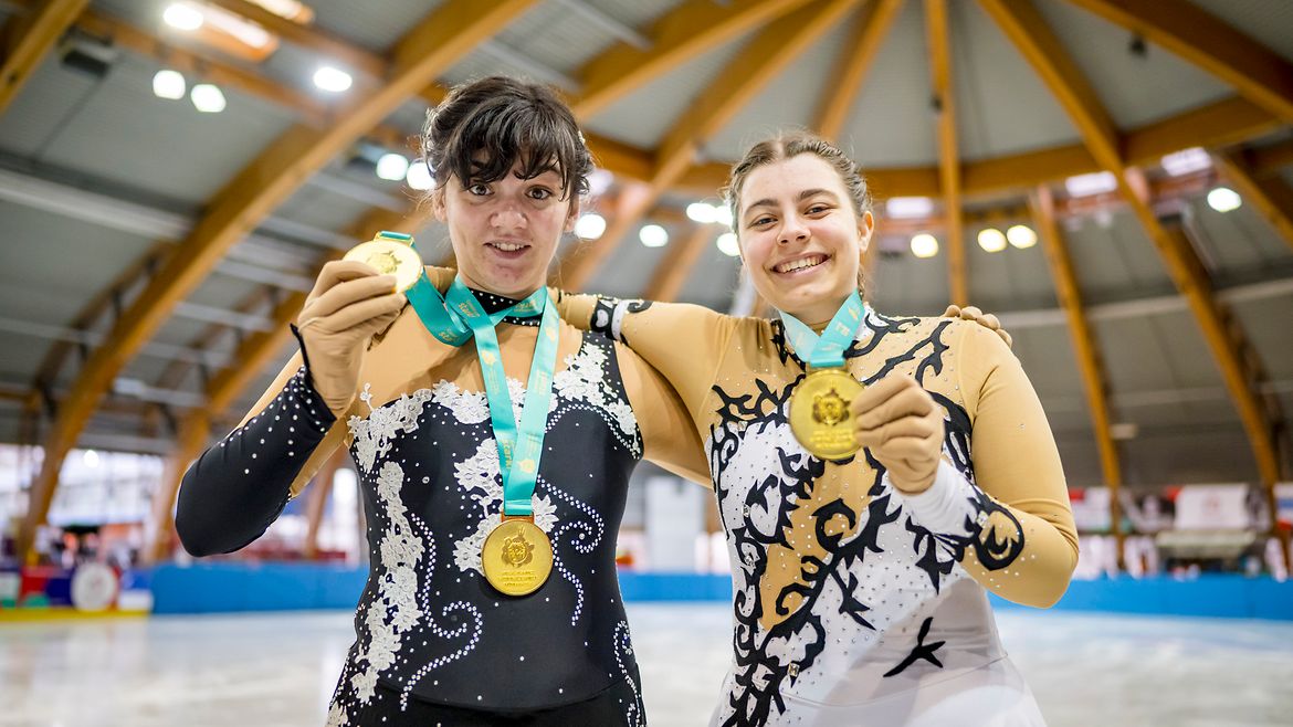Zwei Frauen stehen nebeneinander in einer Eishalle. Beide tragen ärmellose, dekorative Sportbekleidung und halten stolz ihre gewonnenen Medaillen hoch.