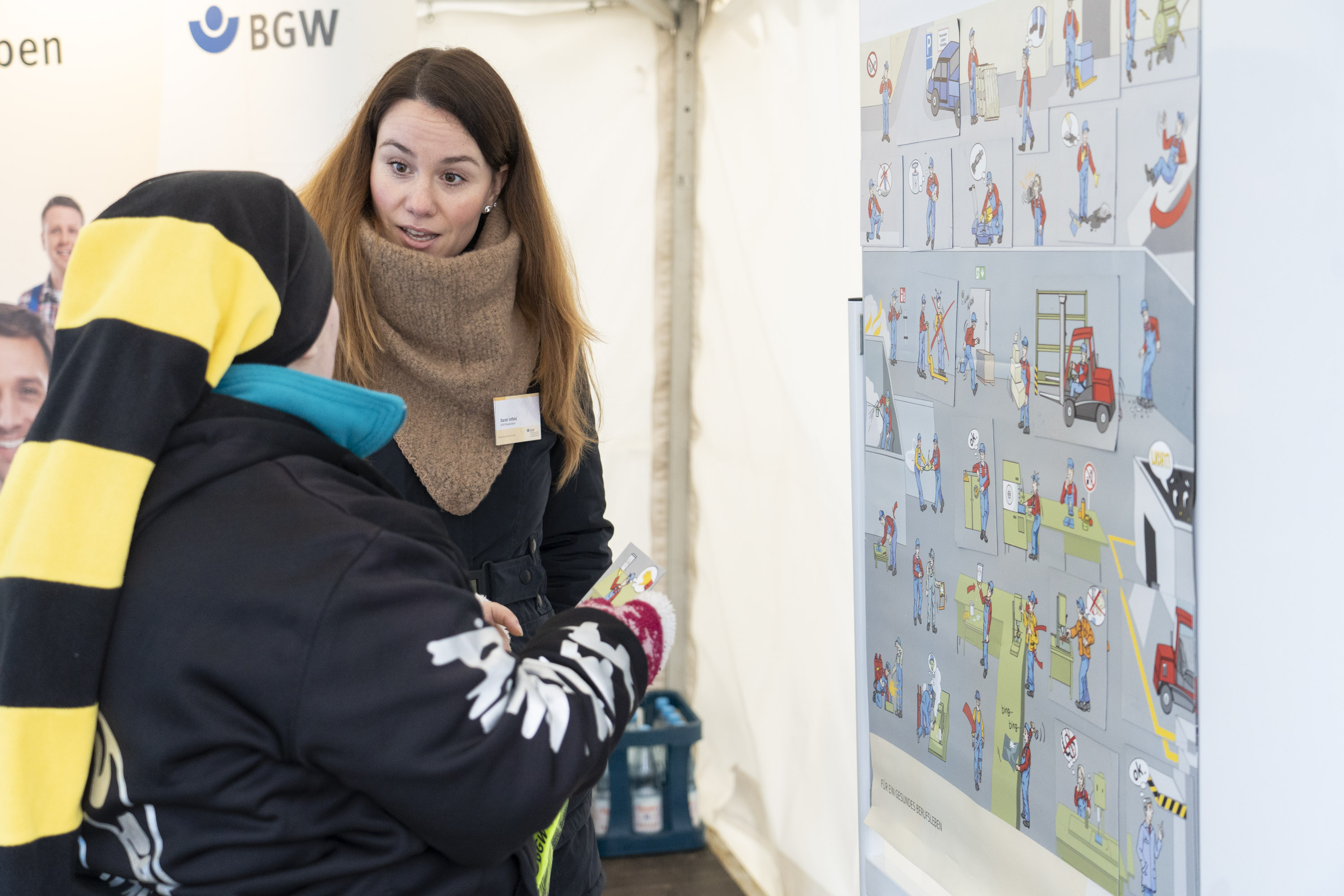 Eine Person in dunkler Jacke und Mütze mit einer Spielkarte in der Hand. Rechts an der Wand ein Plakat mit Illustrationen gefährlicher Situationen in einer Werkstatt. Eine BGW-Mitarbeiterin spricht mit ihr. 