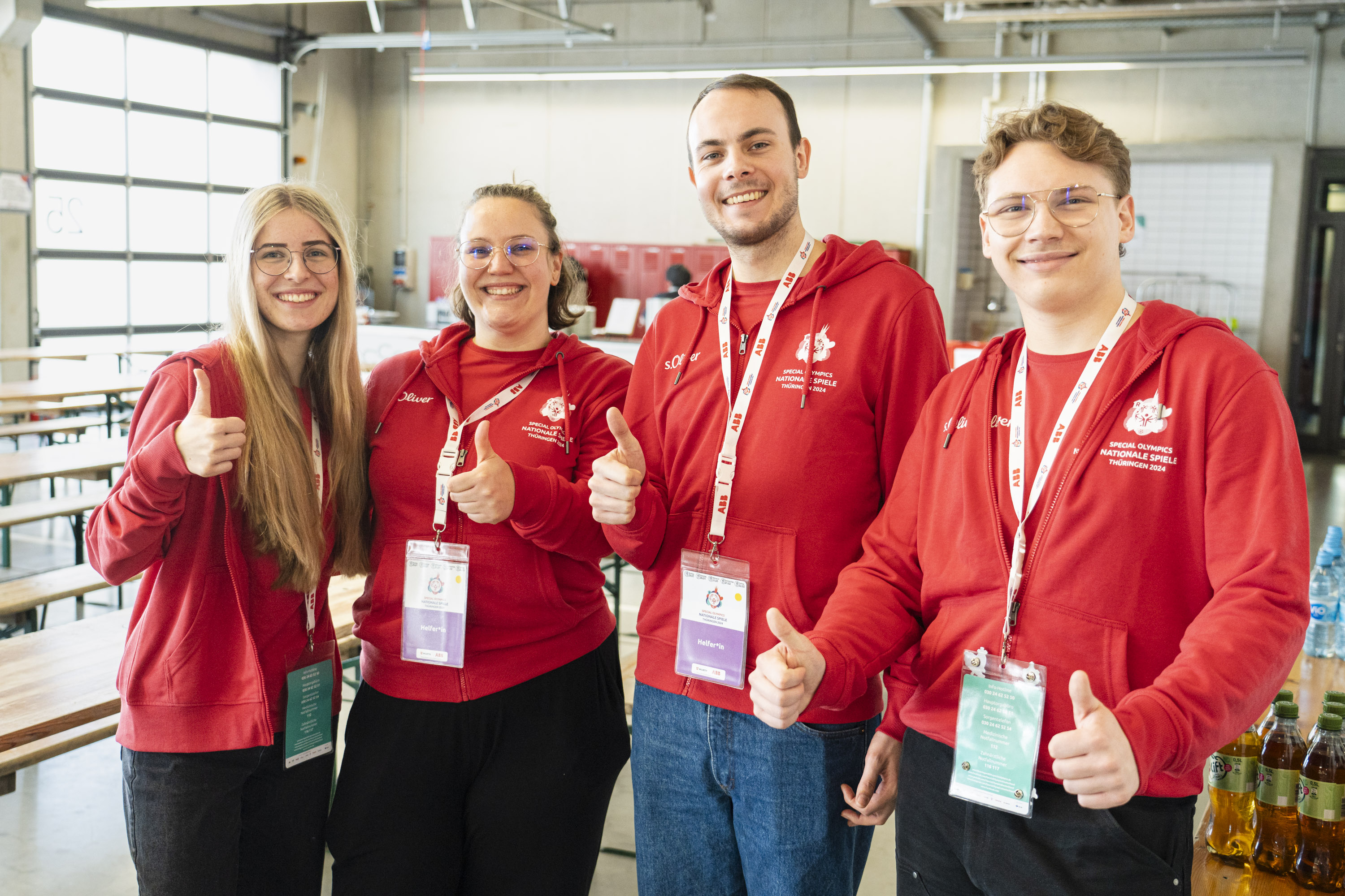 Special Olympics Nationale Spiele Thüringen 2024: Team BGW - Volunteers 2 junge Frauen und zwei junge Männer stehen in roten SOD-Shirts nebeneinander und zeigen alle einen Daumen nach oben. Sie haben alle Bänder mit Anhängern um den Hals, auf denen "Helfende" steht.