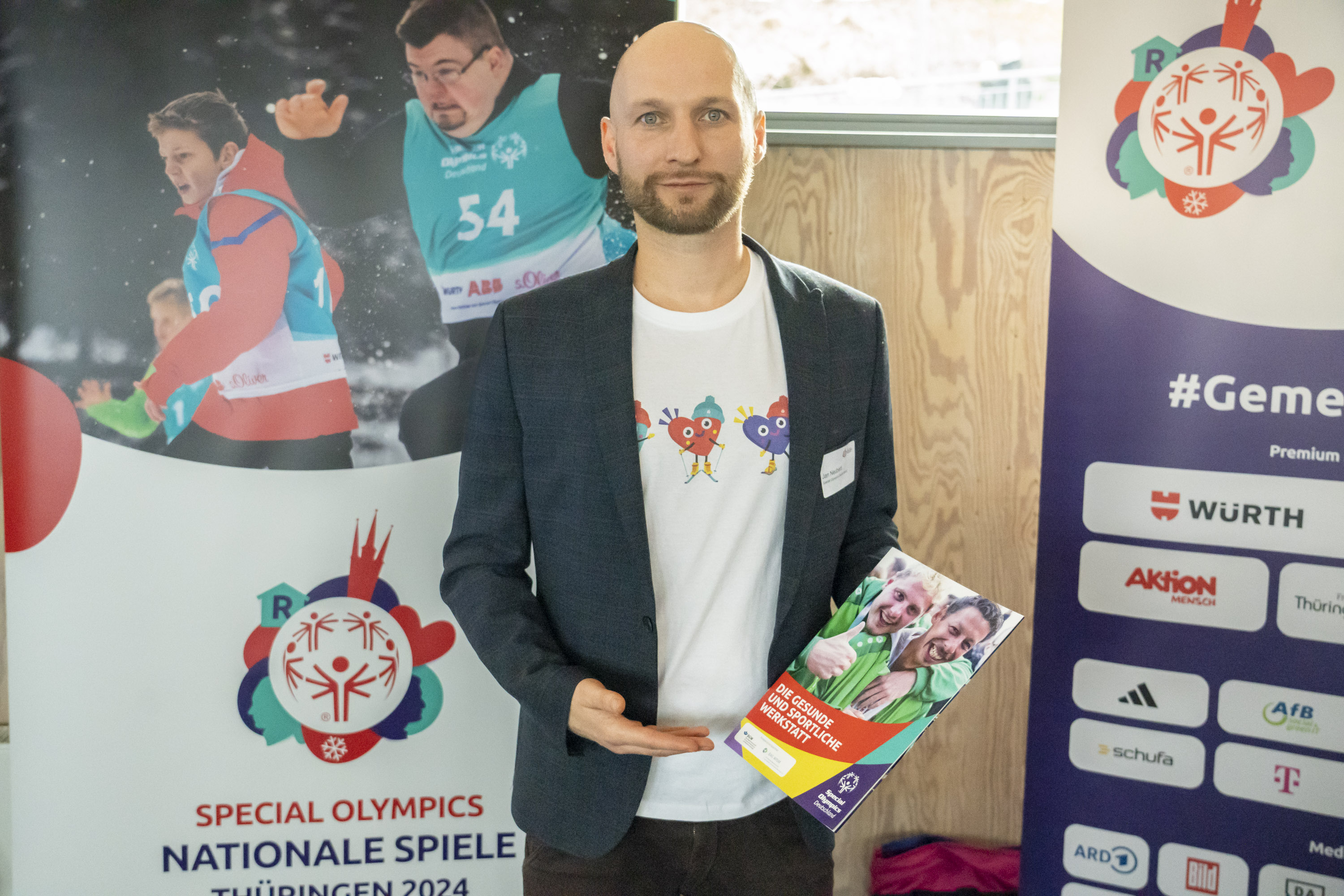 Special Olympics Nationale Spiele Thüringen 2024: Jan Neubert, SOD Eni Mann steht vor 2 Plakaten mit SOD-Motiven undverschiedenen Logos. In der Hand hält er die Broschüre "Die gesunde uund sportliche Werkstatt".