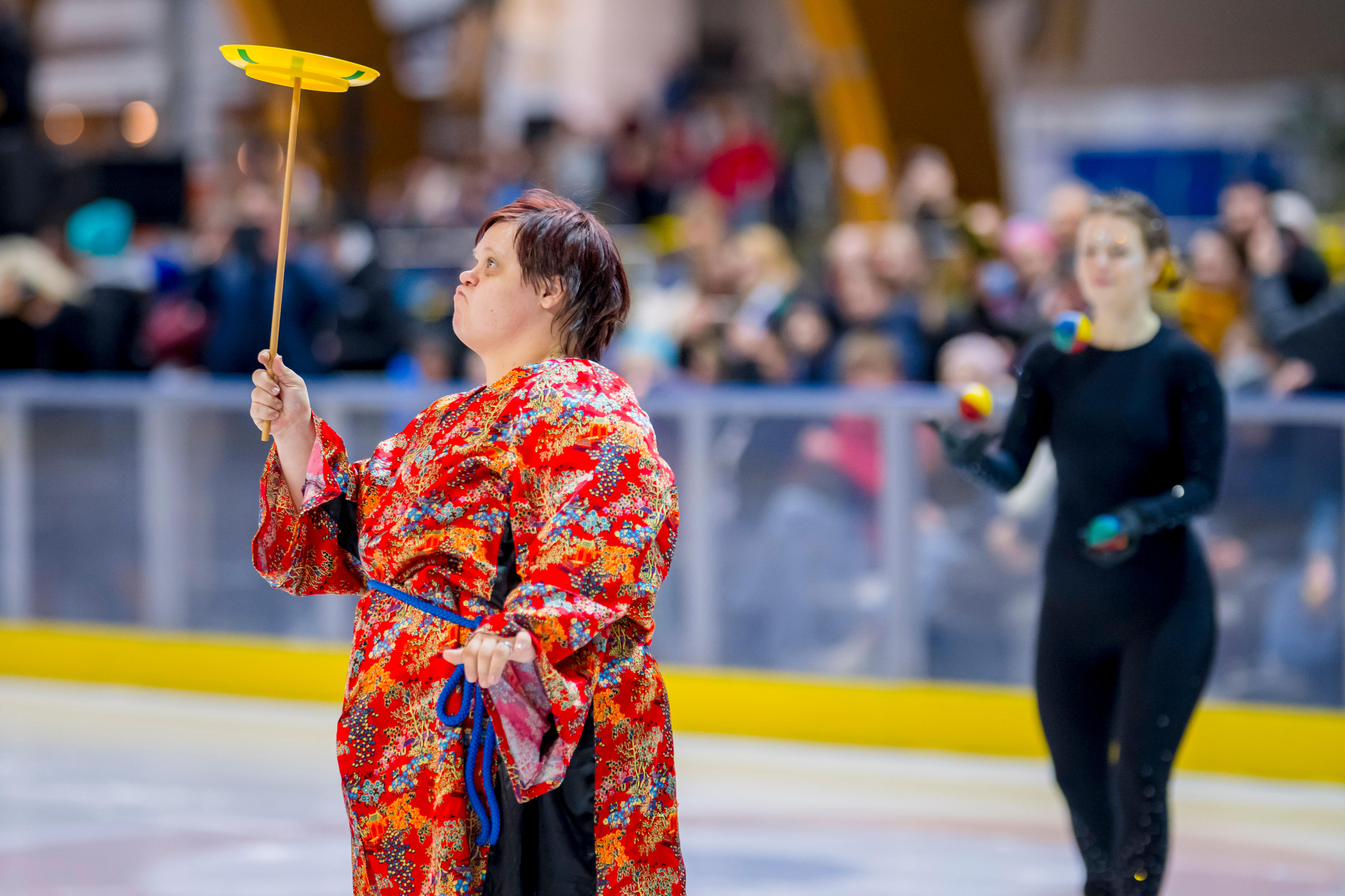 Zwei Athletinnen auf einer Eisfläche im Eisstadion. Eine balanciert einen Teller auf einem Stock, die andere jongliert mit 3 Bällen.