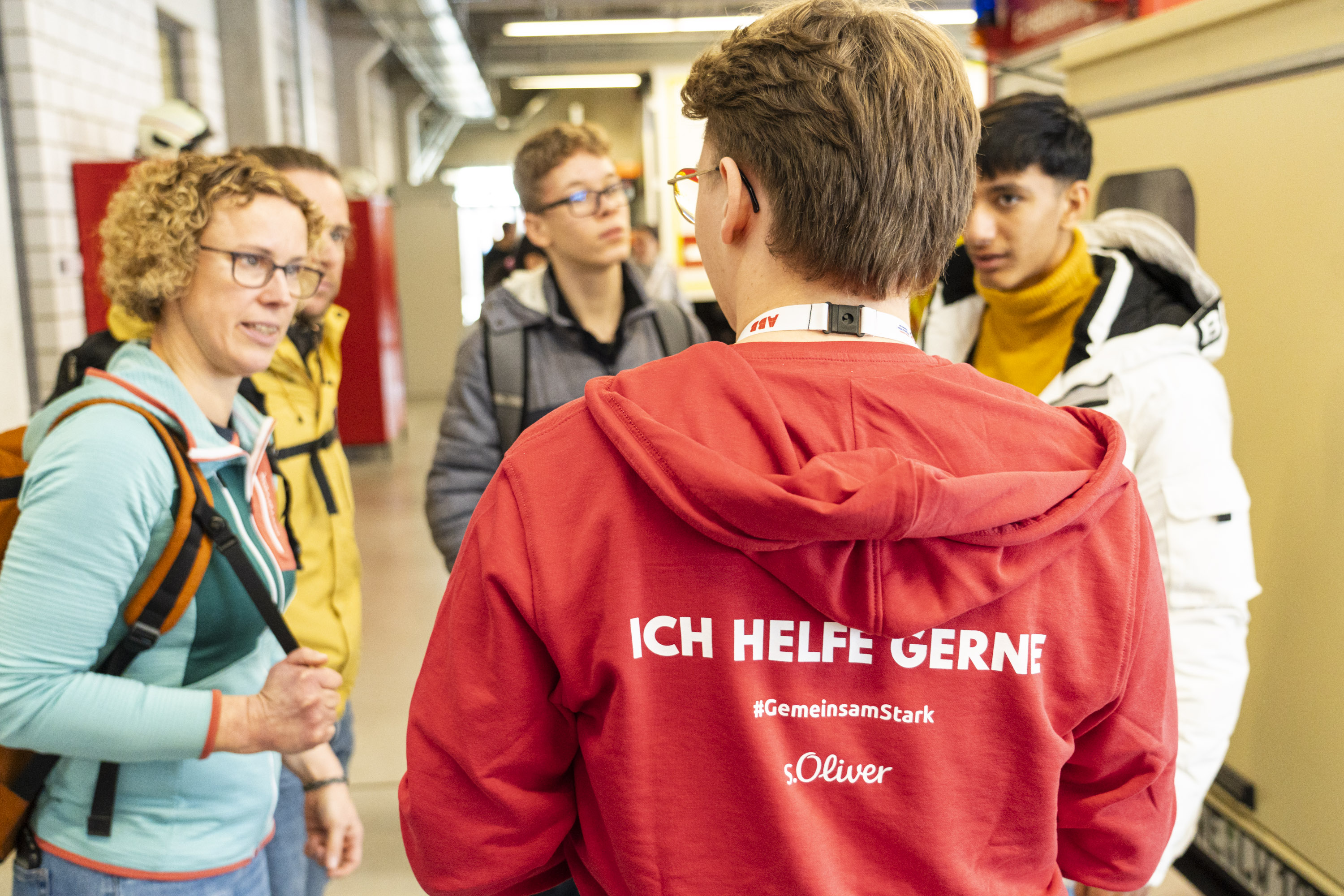 Special Olympics Nationale Spiele Thüringen 2024: Team BGW - Volunteers Ein junger Mann von hinten. Er trägt einen roten Pulli, auf dem steht "Ich helfe gerne!" 4 Personen stehen vor ihm und sprechen mit ihm.