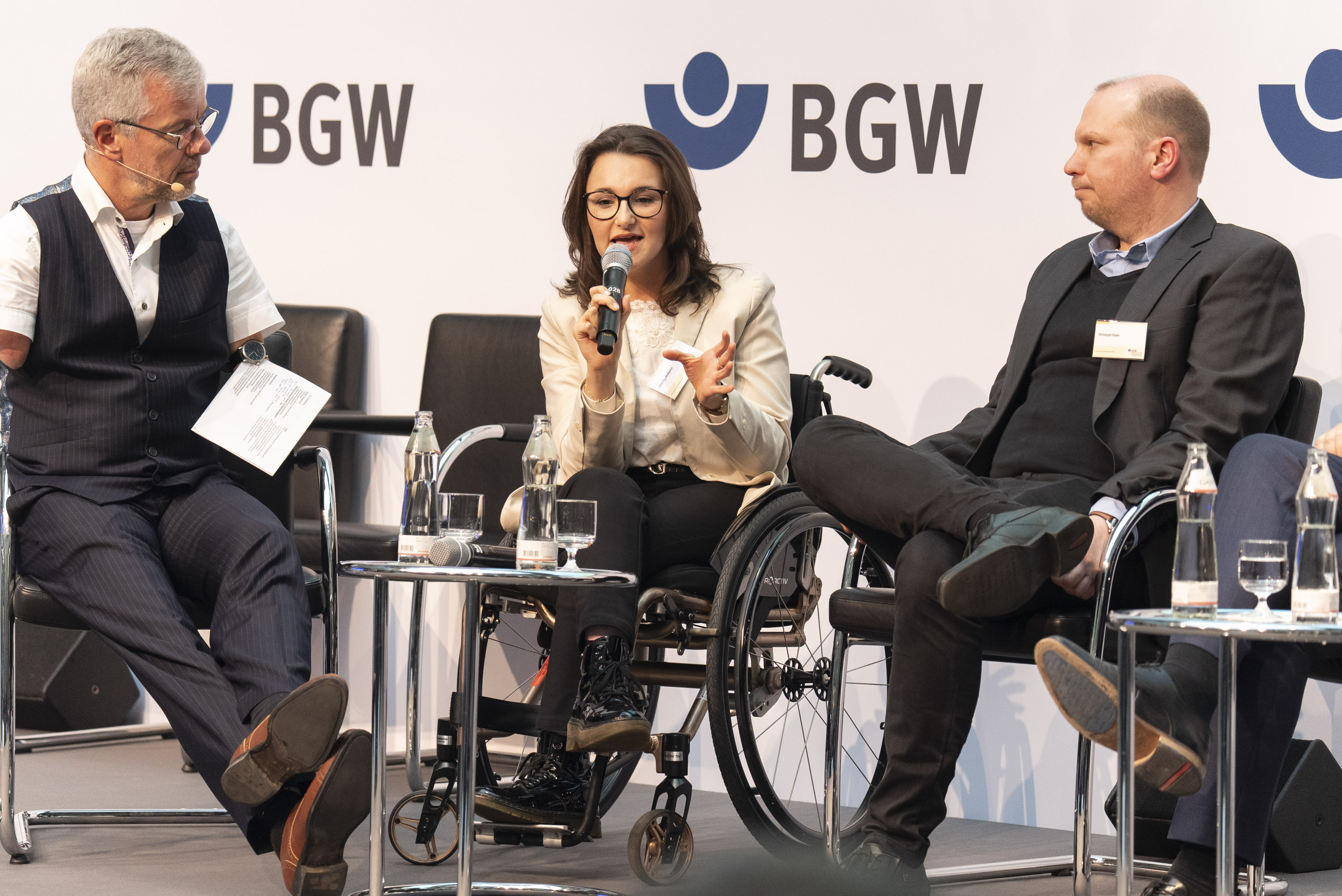 2 Männer und eine Frau auf einem Podium. Der linke hat eine Behinderung an beiden Armen. Die Frau in der Mitte sitzt im Rollstuhl.