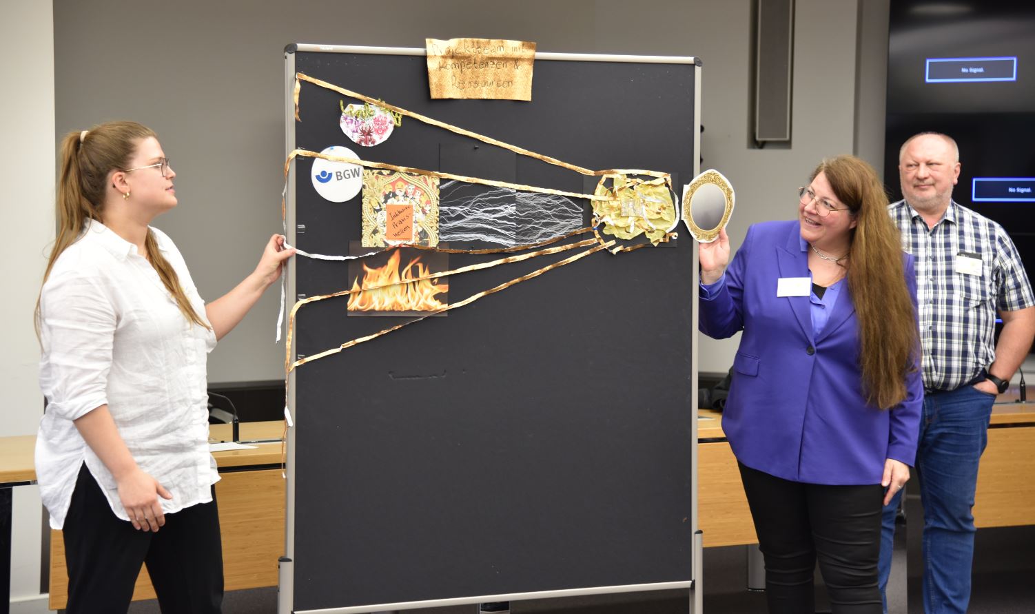 Zwei Damen links und rechts neben einer Stellwand. Die rechte hält das Bild eines Spiegels an die Stellwand. Über die Stellwand ziehen goldene Strahlen in Richtung verschiedener Symbole (BGW Logo, Feuer ...).