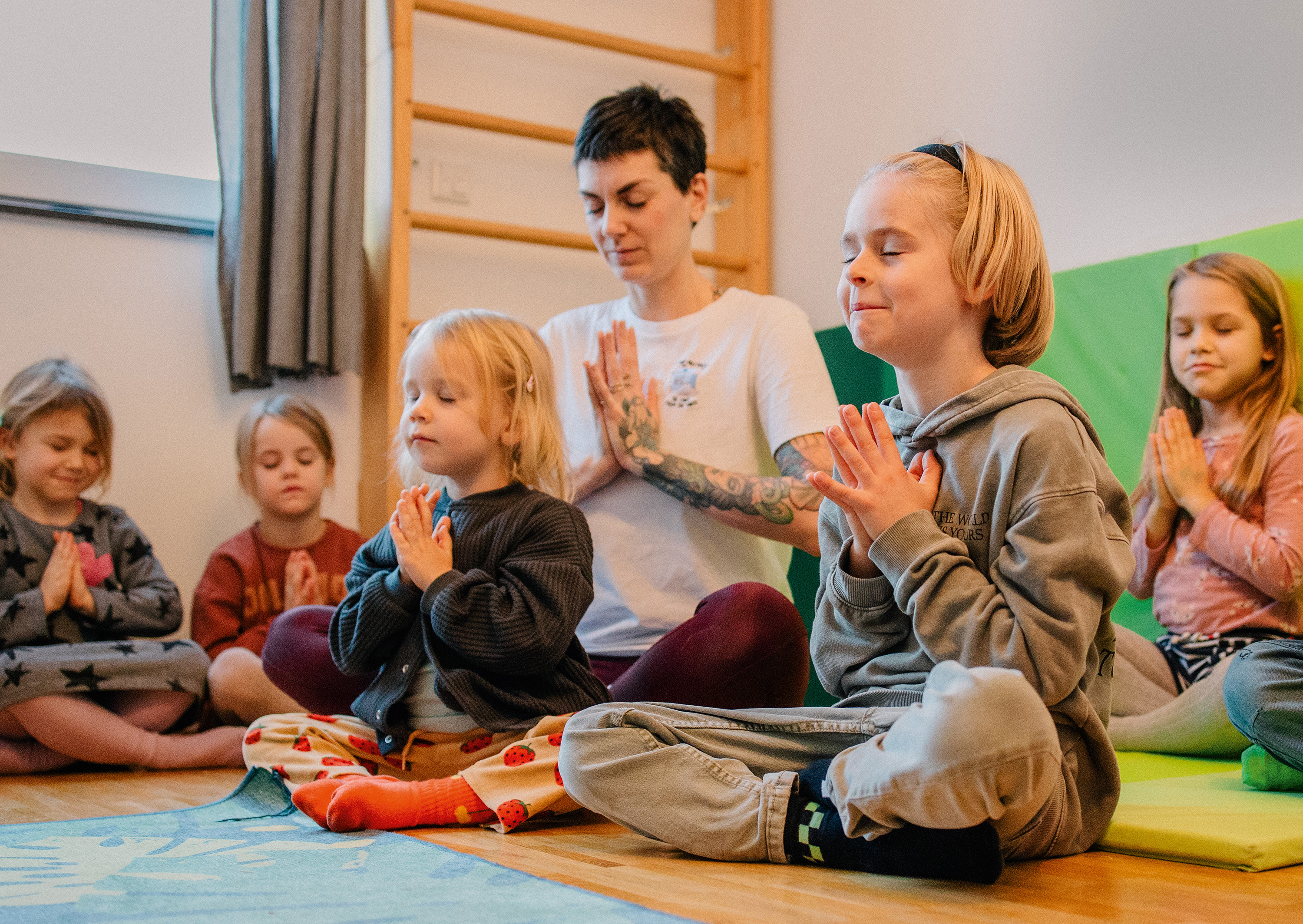 Beatrice Hong "Mitte stärken – Zukunft stärken" Eine Frau und mehrere Kinder um sie herum sitzen im Schneidersitz auf dem Boden, haben die Hände vor der Brust gefaltet und die Augen geschlossen