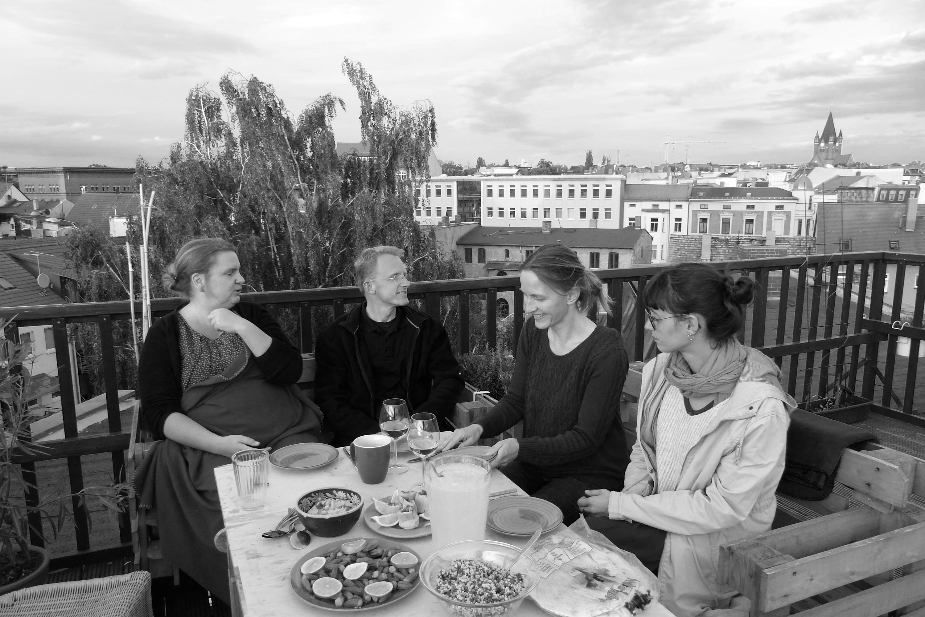 Ellen Schüttig "Vitamin B und N – Beziehungen und Natur" Schwarz-weiß-Aufnahme: Ein Mann und drei Frauen sitzen auf einer Terasse an einem gedeckten Tisch, im Hintergrund ist ein Stadtpanorama zu sehen.