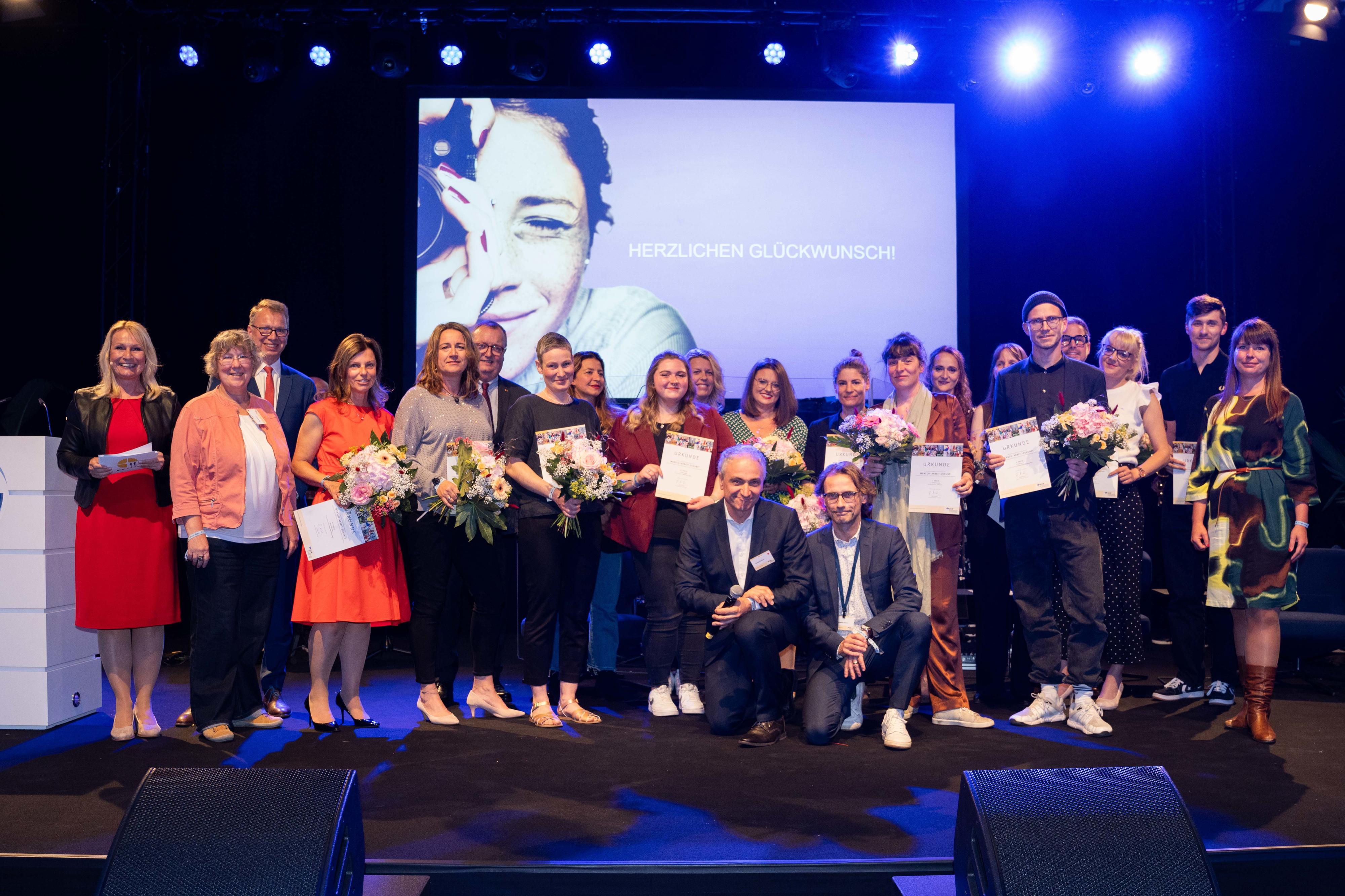 Alle Gewinner des Fotowettbewerbs mit Urkunde und Blumenstrauß in Händen zusammen mit Mitgliedern der Jury auf einer Bühne.