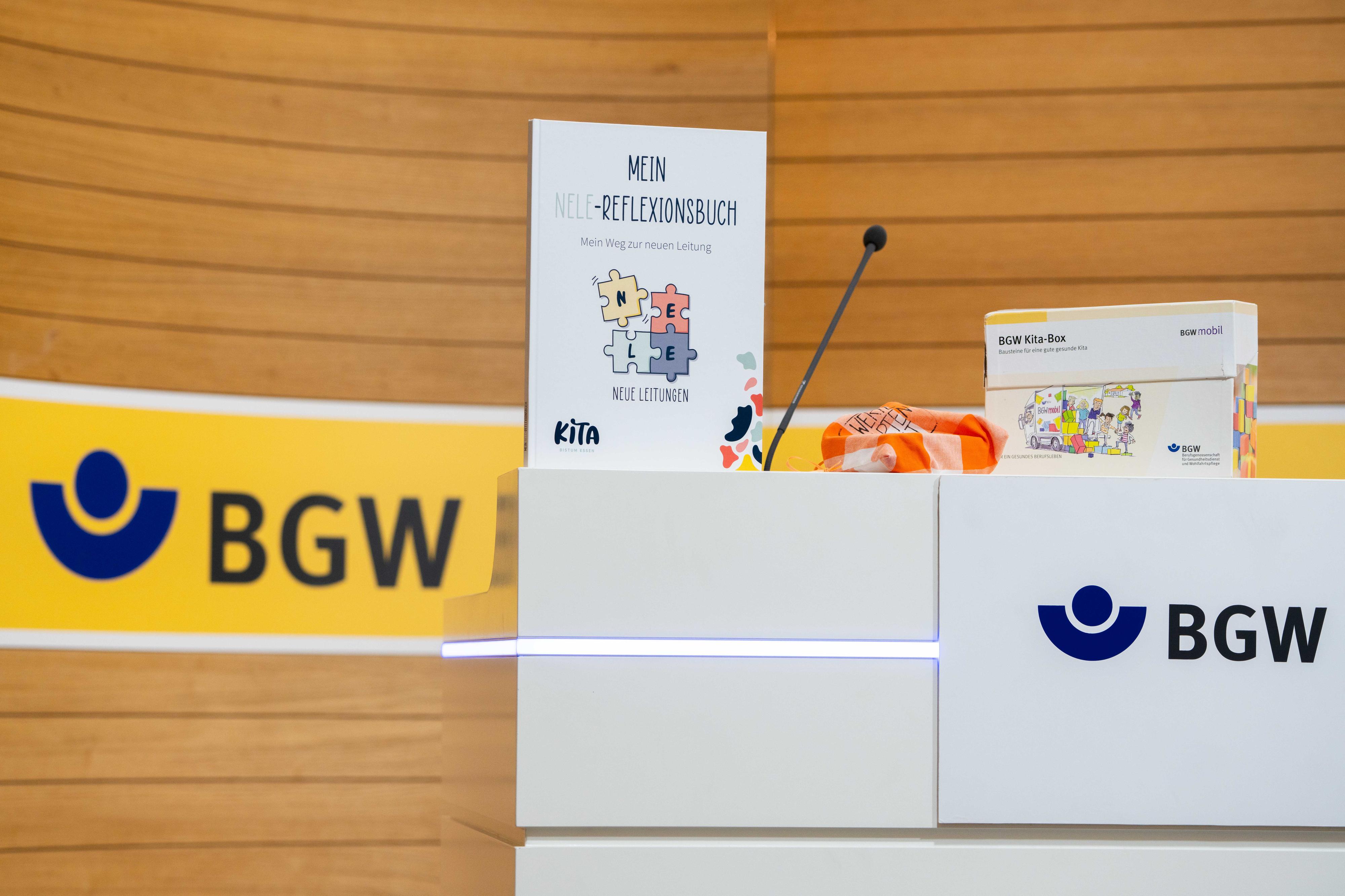 Auf einem Tisch mit dem Logo-Schriftzug "BGW" stehen unter anderem ein Karton mit der Aufschrift "BGW Kita-Box" und ein Aufsteller mit Überschrift "Mein Nele-Reflexionsbuch".