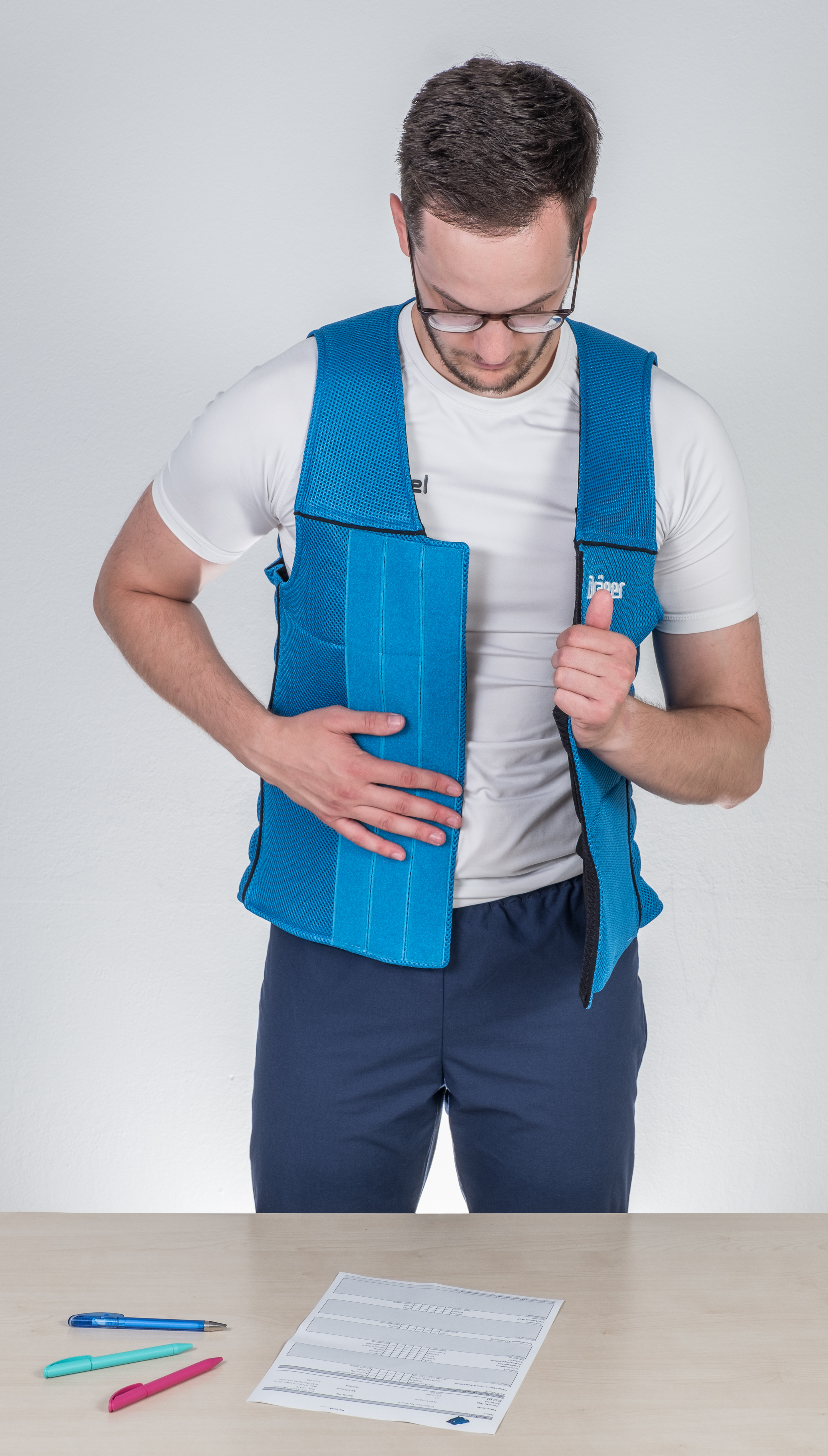 BGW test Kühlwesten in der Anwendung Ein Mann zieht eine blaue Kühlweste über sein Shirt. Vor ihm auf dem Tisch liegt ein Fragebogen mit Stiften.