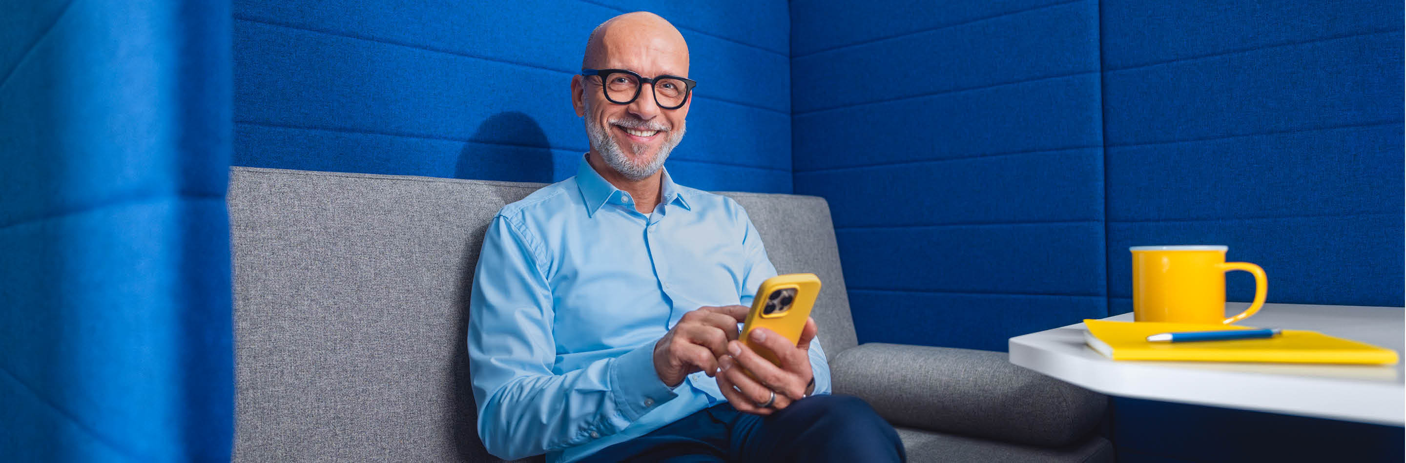 1000 UNDEIN GRUND – Deine Idee von Karriere: mehr bewirken. Ein Mann sitzt auf einem Sofa vor einer blauen Wand und hält ein Smartphone in der Hand.