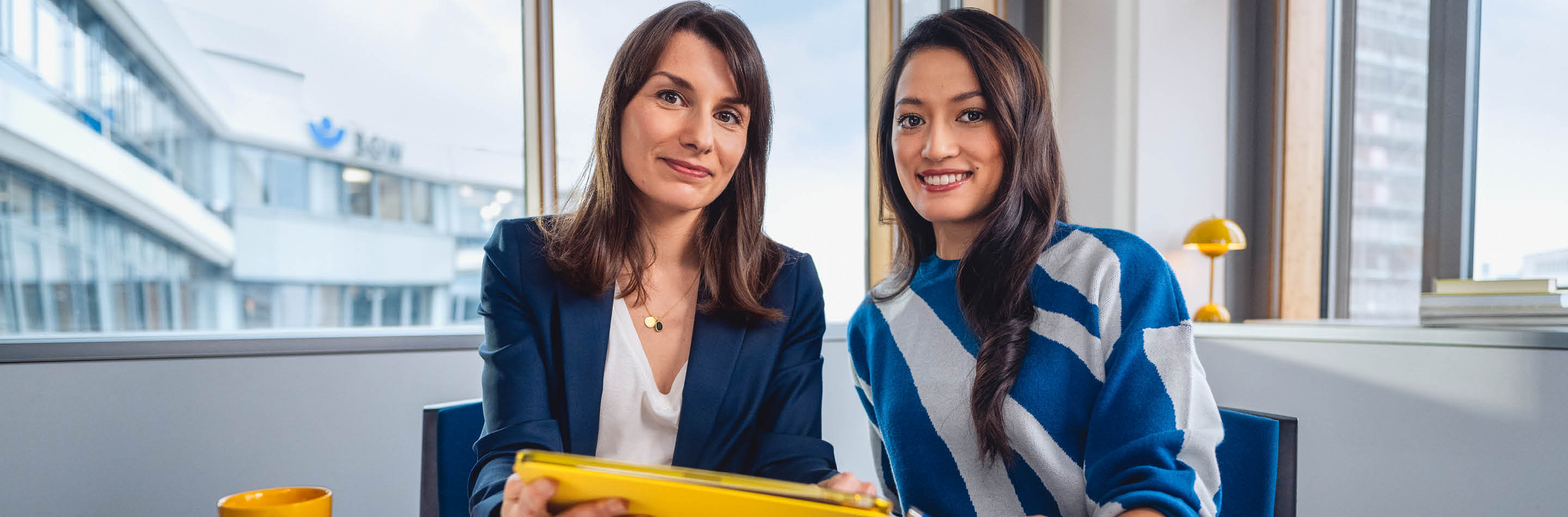 1000 UNDEIN GRUND – Deine Idee von Karriere: mehr bewirken. Zwei junge Frauen sitzen nebeneinander vor einem Fenster im Büro und lächeln in die Kamera.