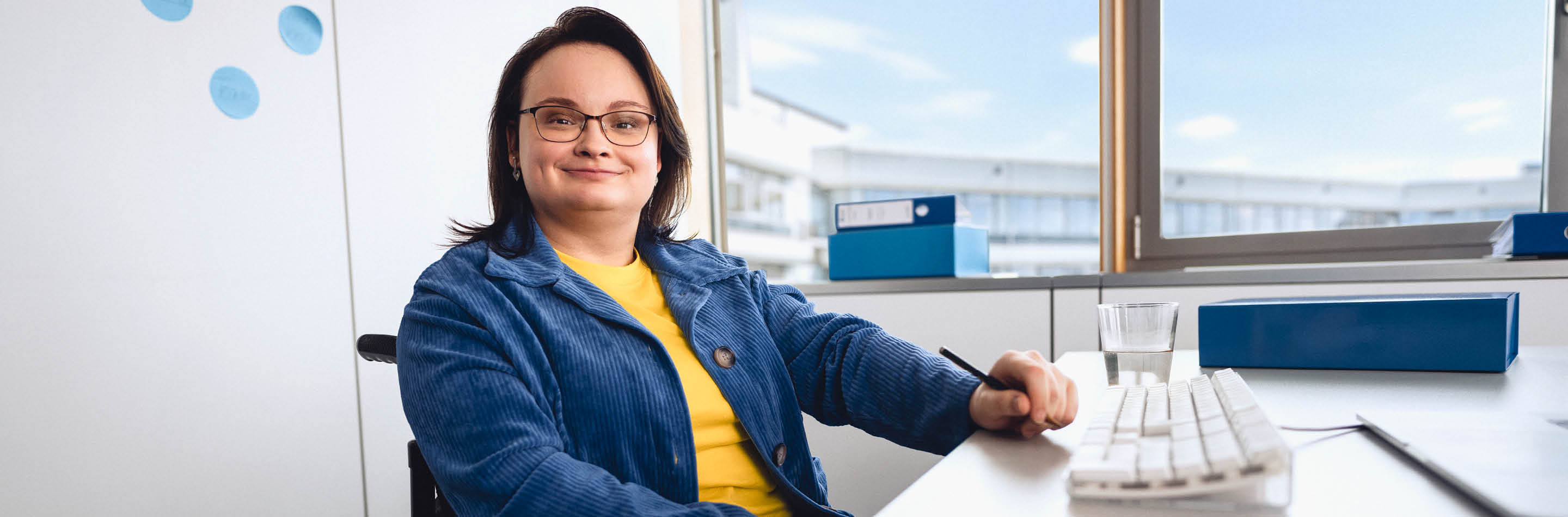 Eine Frau im Rollstuhl sitzt am Schreibtisch in einem Büro.