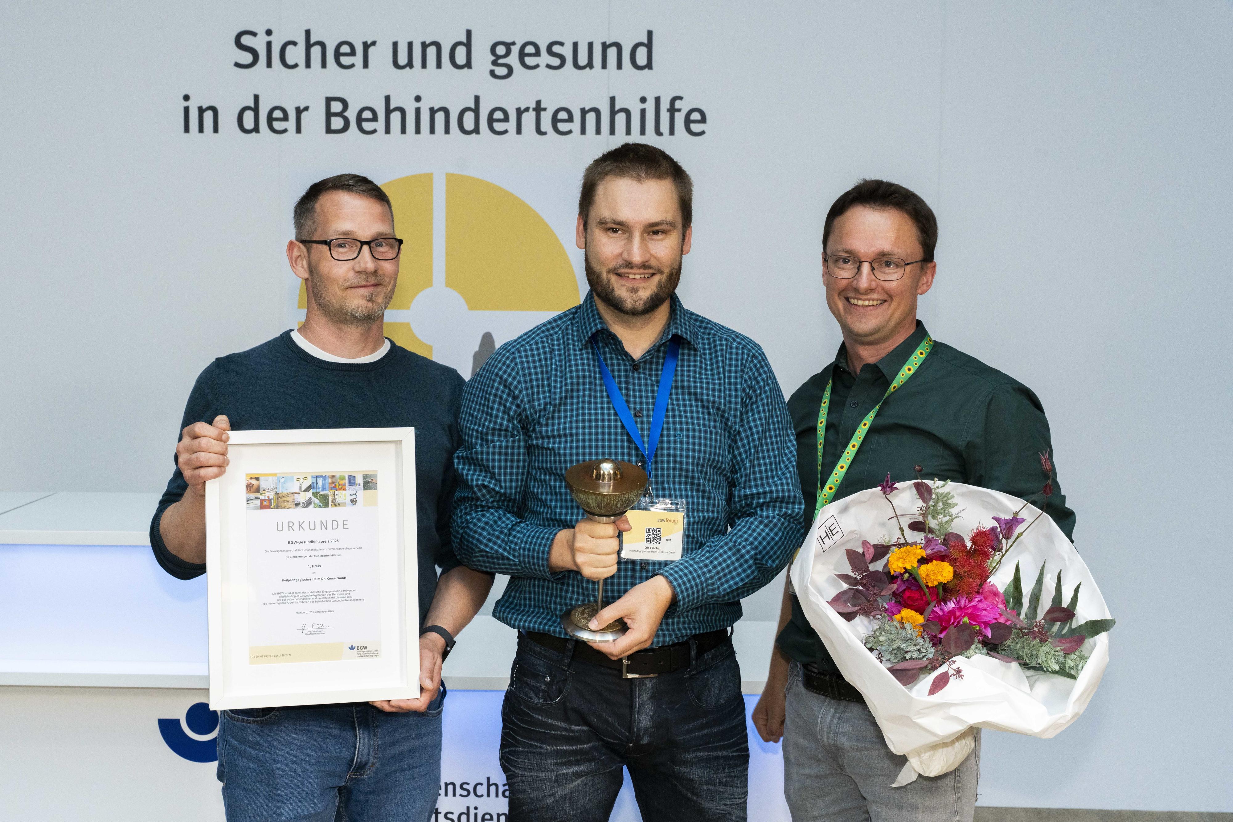 Drei lächelnde Personen, eine davon mit Blumenstrauß, eine weitere mit Urkunde.