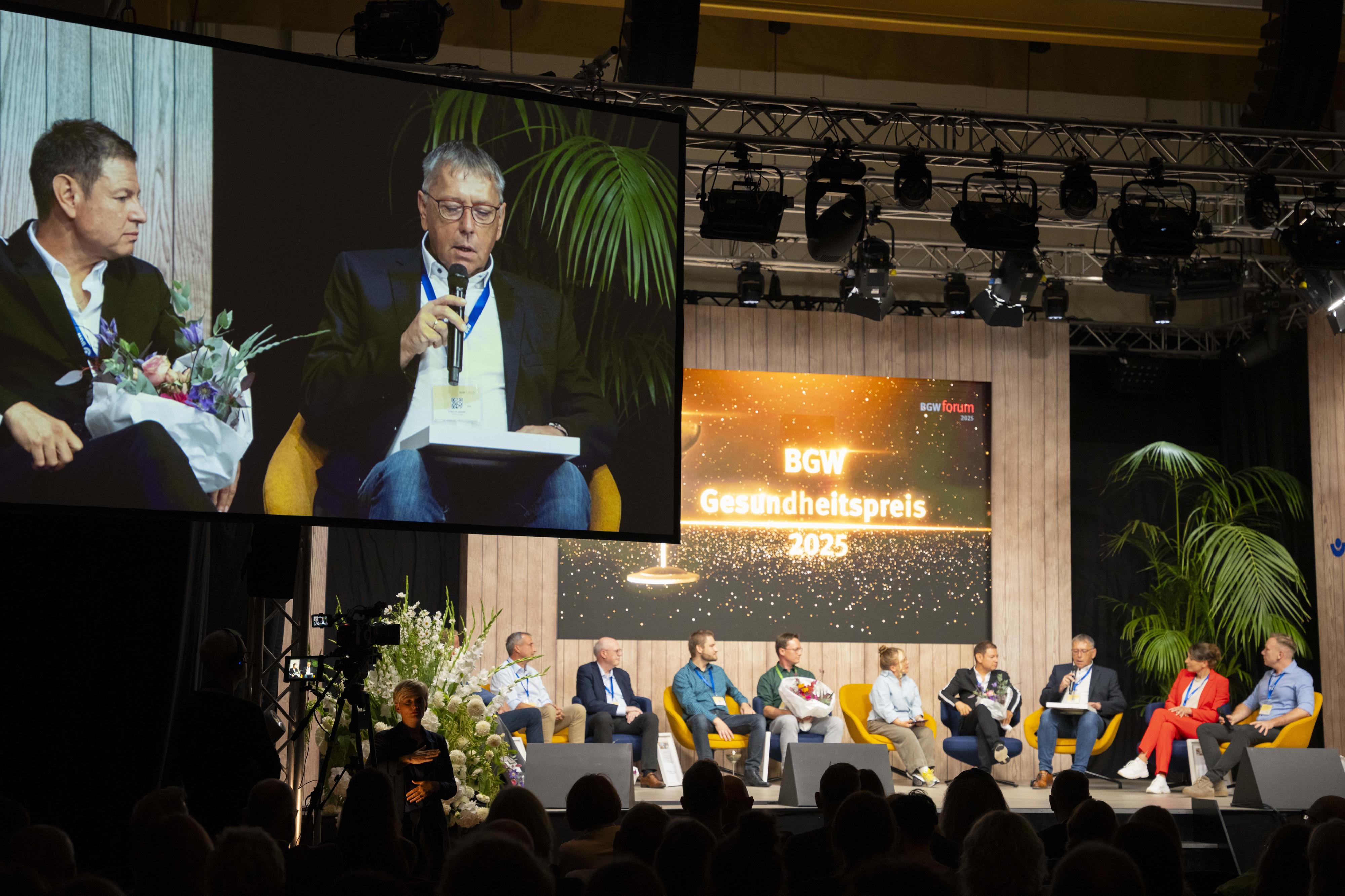 Mehrere Personen auf einer Bühne im Gespräch, im Hintergrund eine leuchtende Leinwand mit der Aufschrift "BGW Gesundheitspreis 2025"